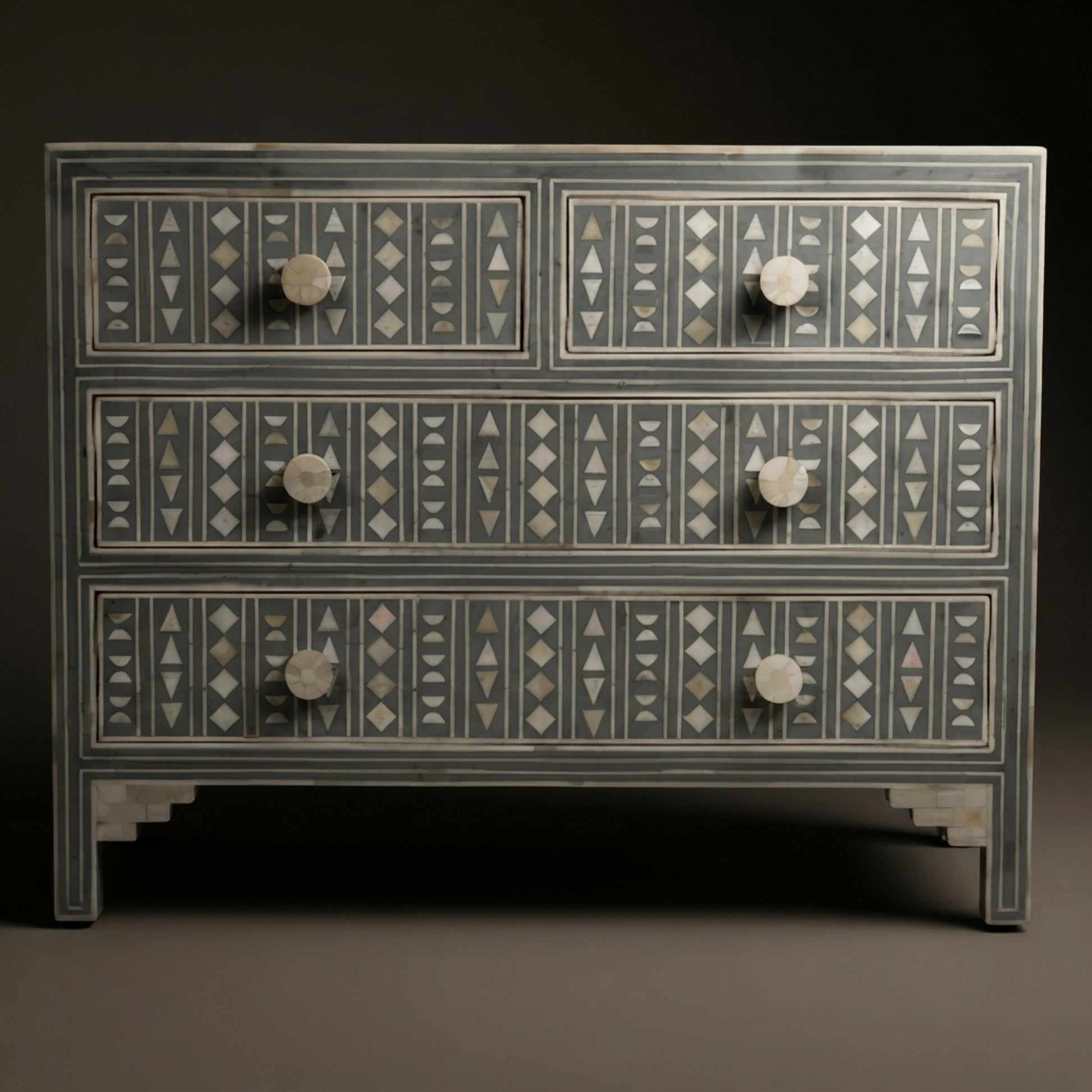 A three-drawer dresser with a dark gray painted finish and intricate white and gray geometric inlay patterns on the drawer fronts, featuring round white knobs, set against a dark background.