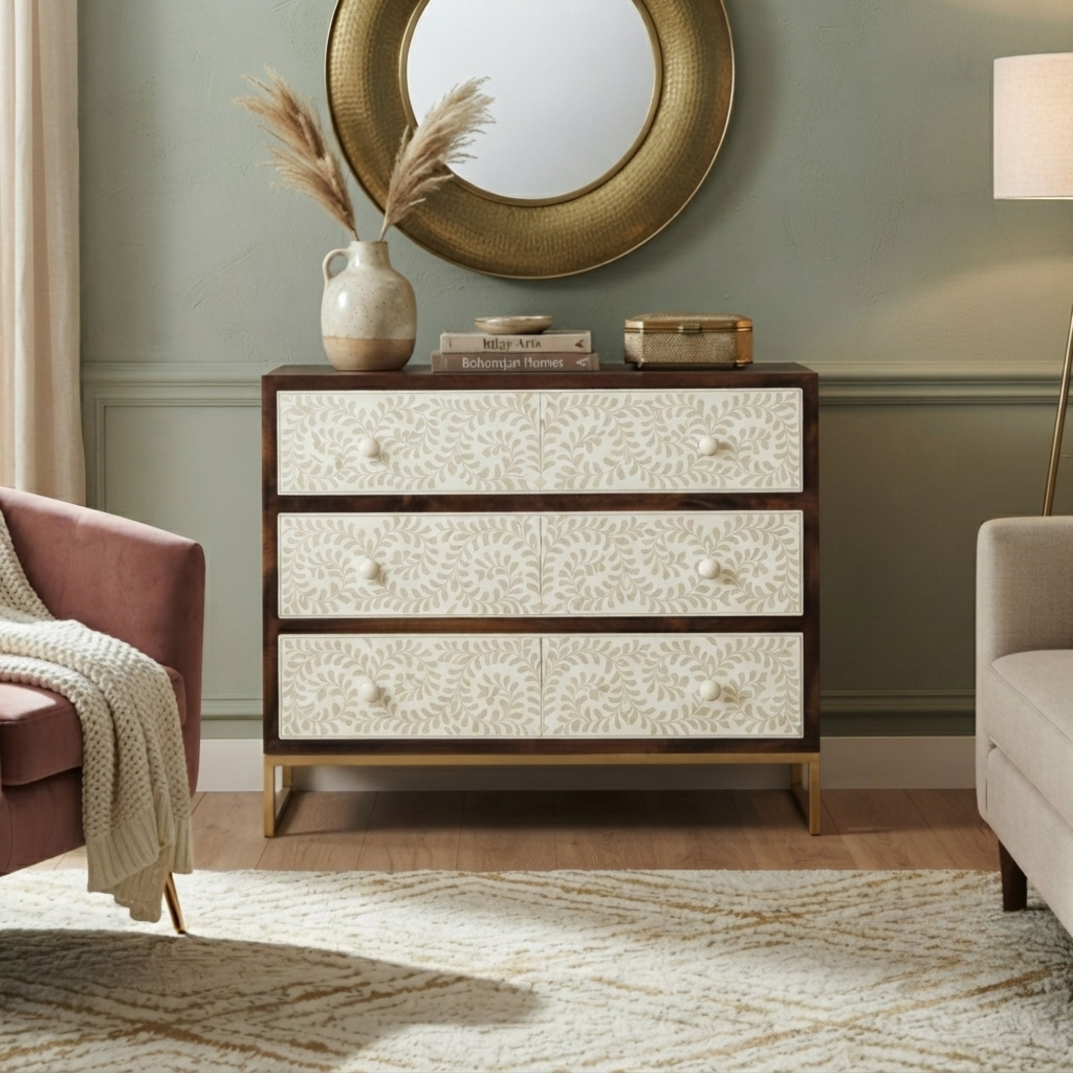 A three-drawer chest of drawers in a styled living room setting, featuring cream floral-patterned fronts, dark brown wood trim, and gold metal legs, topped with a vase of pampas grass and books.
