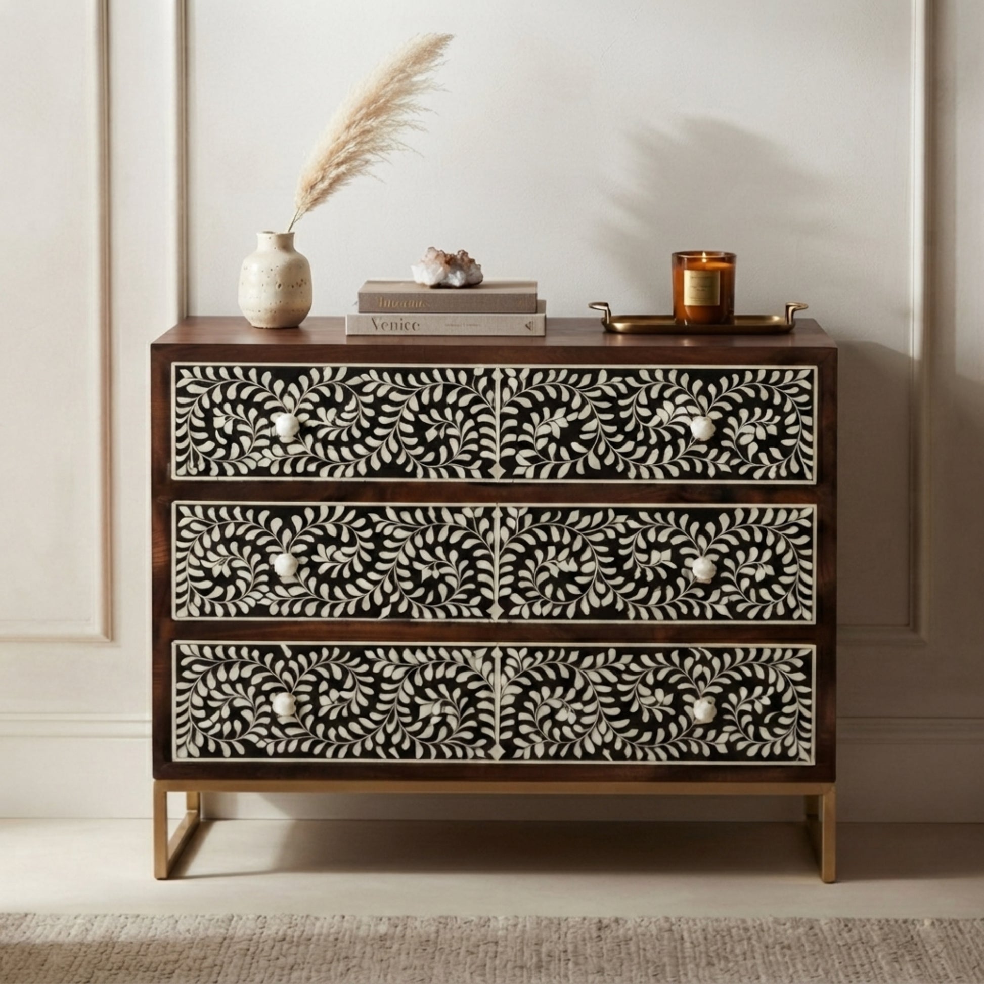 A three-drawer chest of drawers with a dark wood frame and intricate black-and-white floral bone or shell inlay on the drawer fronts, styled with decorative objects on top against a light paneled wall.