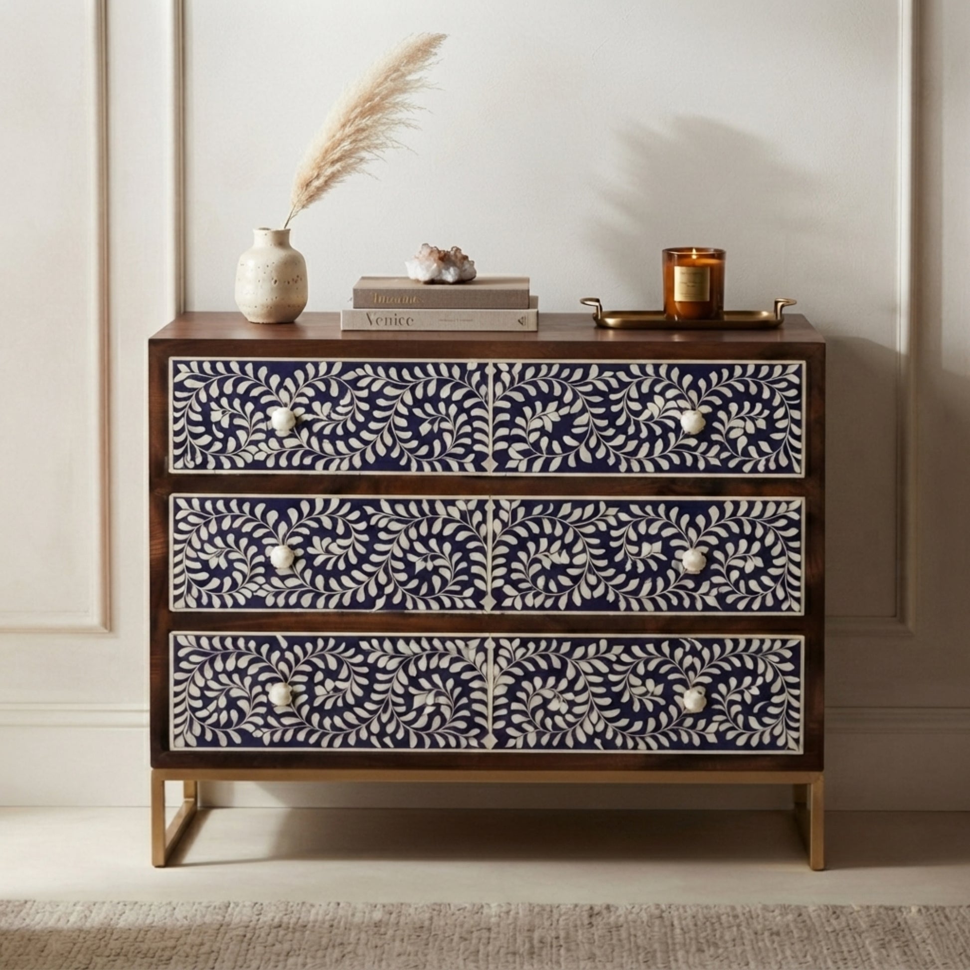 A three-drawer chest of drawers with dark wood frames and intricate white floral bone or shell inlay on the drawer fronts, styled with home decor items on top against a neutral paneled wall.