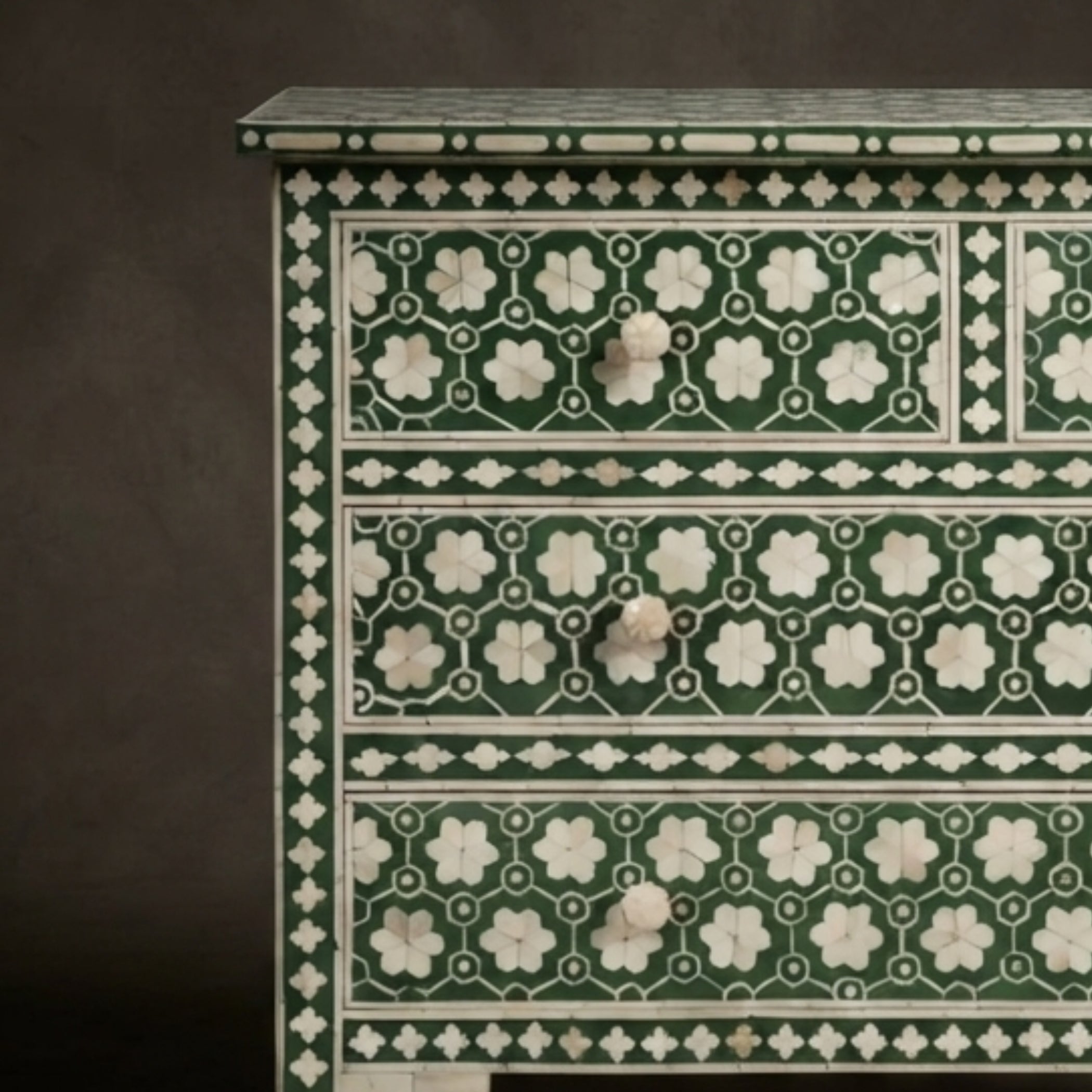 A close-up of an ornate chest of drawers featuring dark green and white floral inlay patterns on the front panels, with three visible drawers and round drawer pulls, set against a dark brown background.