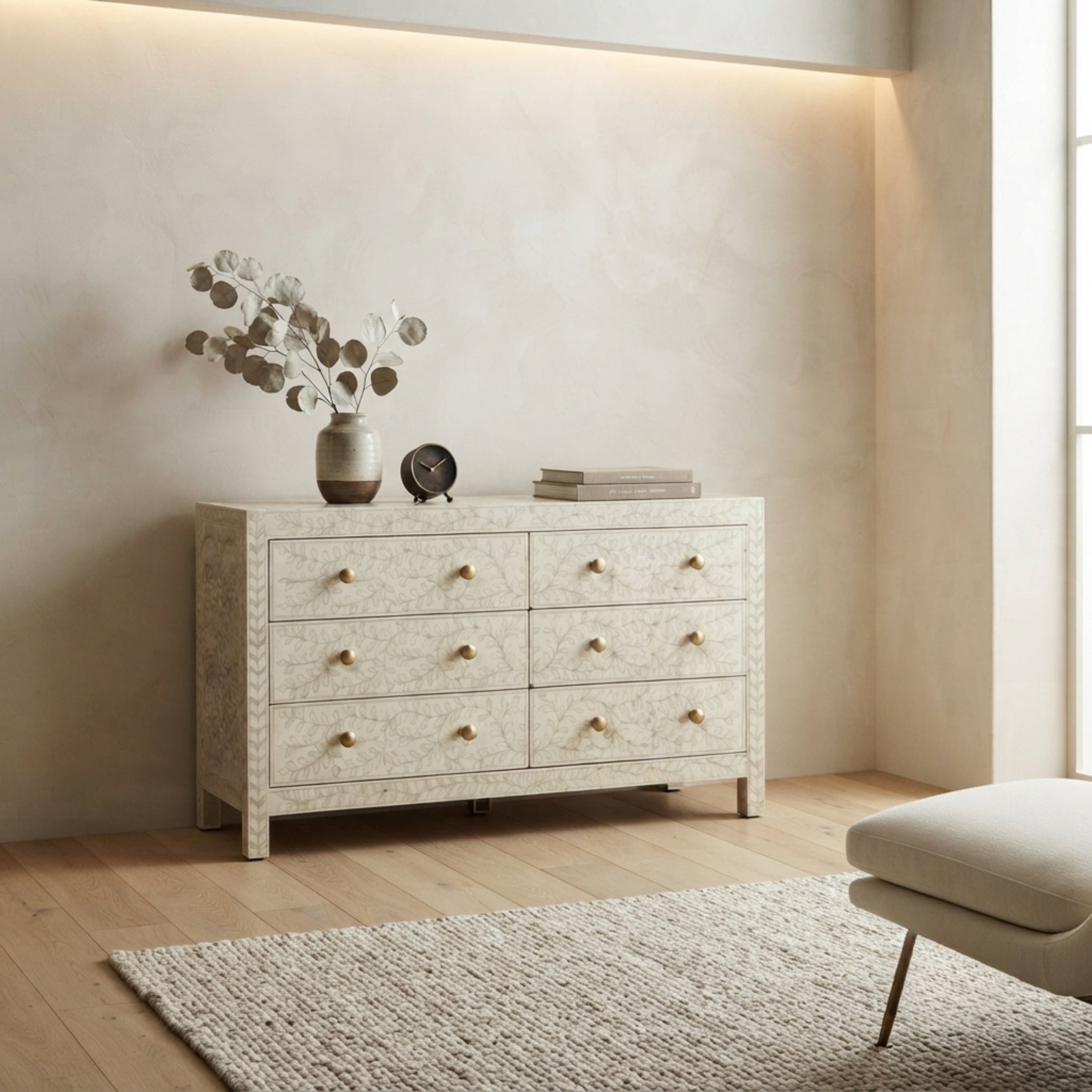 An off-white carved wooden chest of drawers with six drawers and small gold knobs, styled in a warm, modern room with a beige wall, light wood floor, and a matching ottoman.