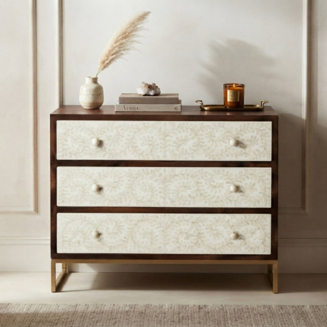 A three-drawer chest of drawers with a dark wood frame, cream patterned drawer fronts, and gold legs, styled with home decor items on top against a neutral paneled wall.