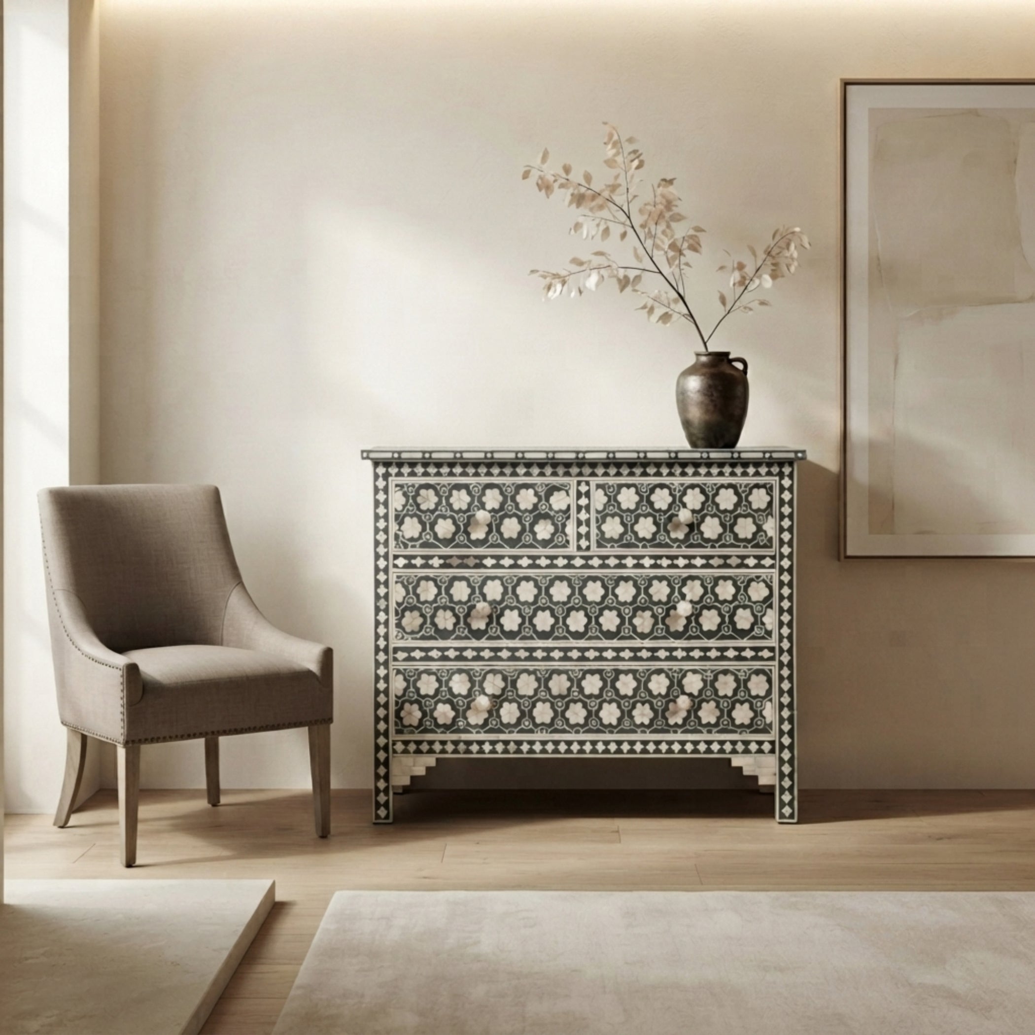 A decorative chest of drawers with an intricate black and white floral bone-inlay pattern, styled in a warm beige room next to a gray armchair with a vase of dried branches on top.