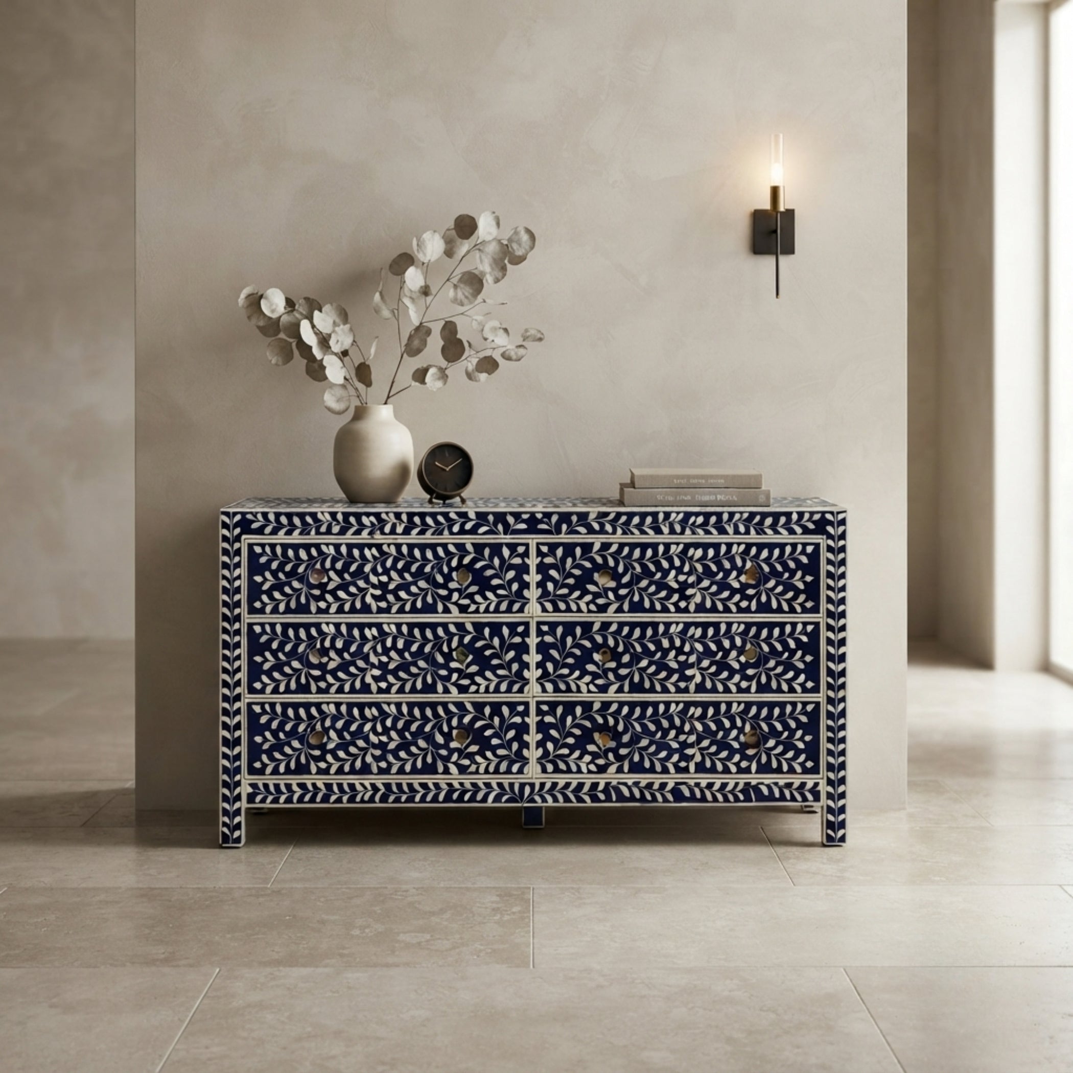 A navy blue and white patterned sideboard or dresser with multiple drawers, styled in a minimalist room with a vase of dried leaves, books, and a wall sconce.