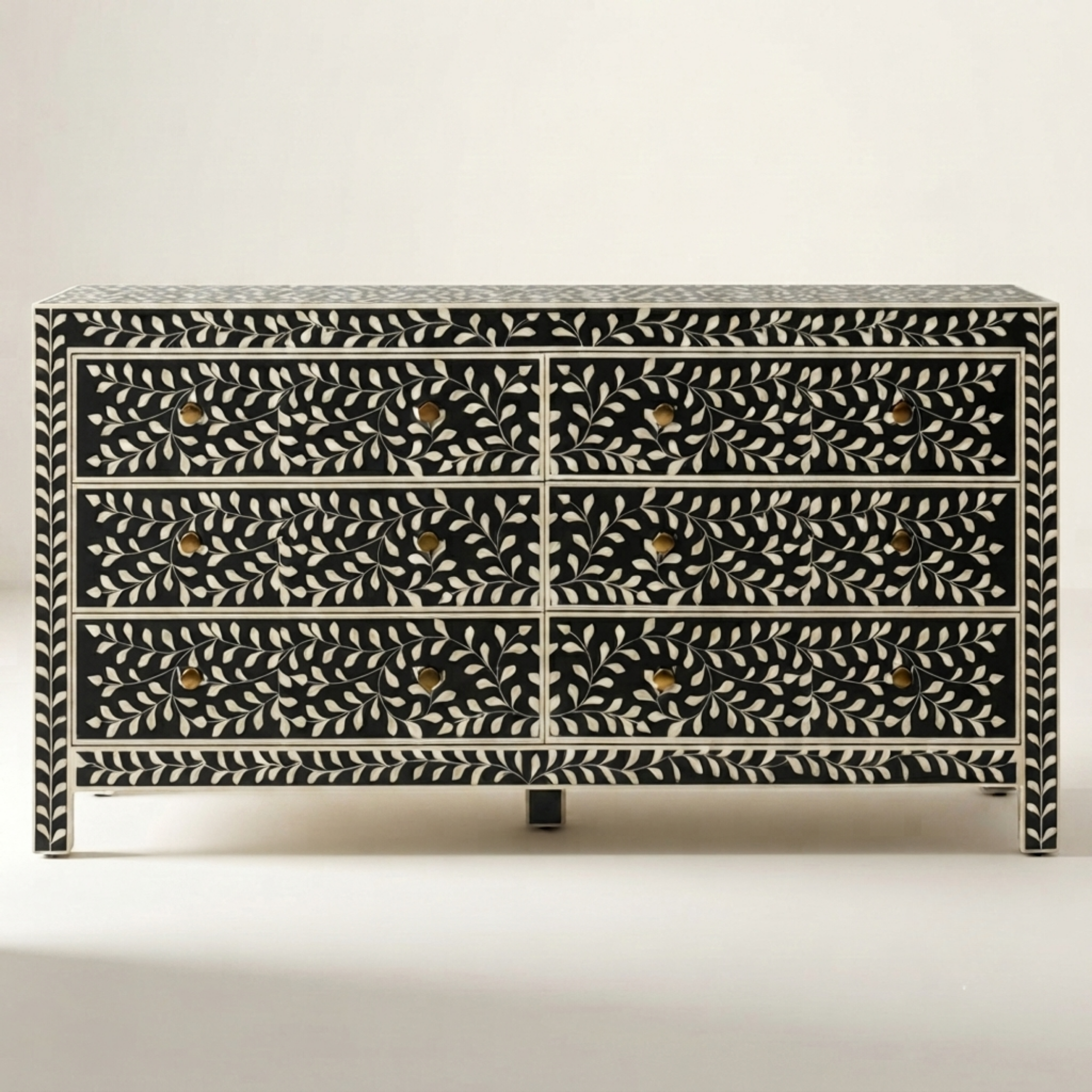 A bone-in-bone chest of drawers with a black and white leaf pattern and small gold knobs, photographed against a neutral light gray background.