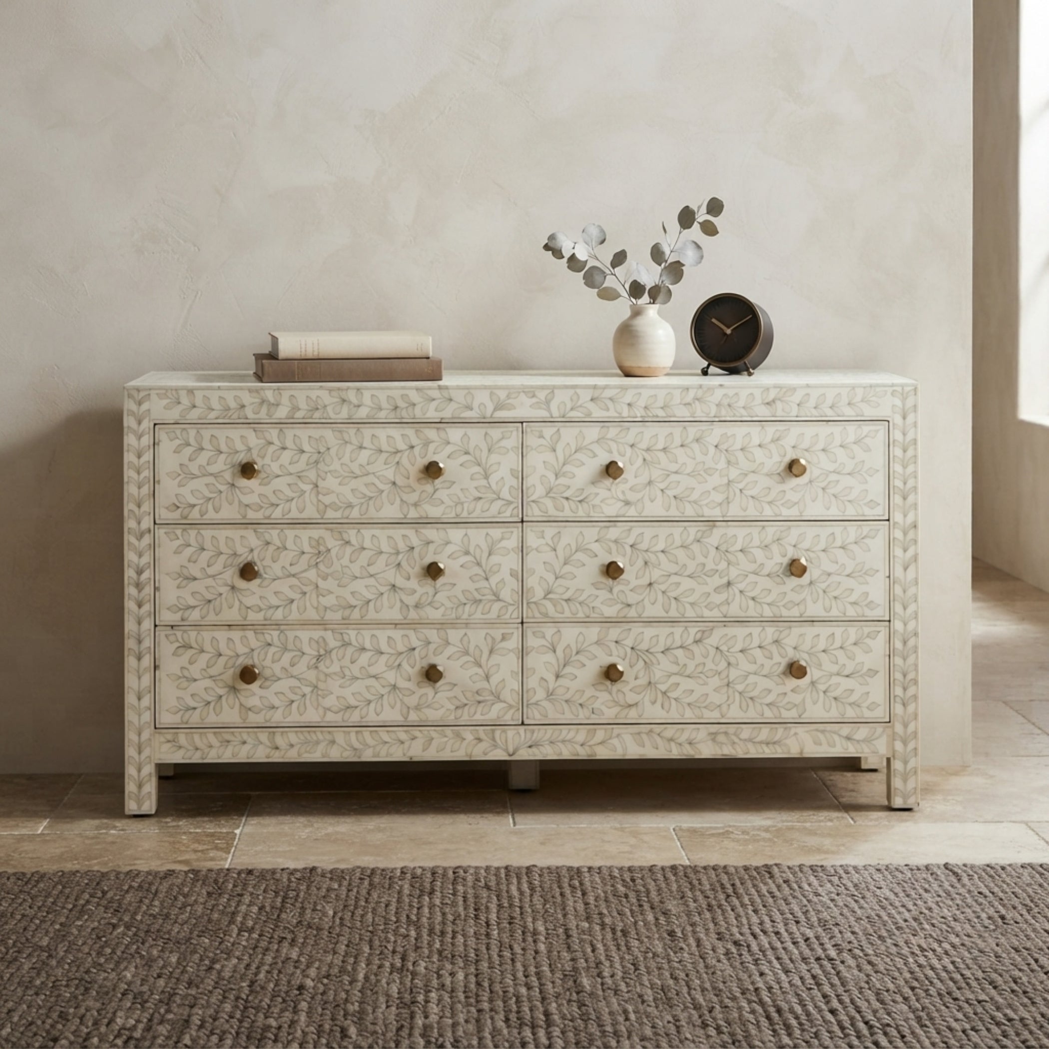 An eight-drawer dresser with an embossed floral pattern and brass knobs, styled in a neutral-toned room with decorative books, a vase, and a clock on top.