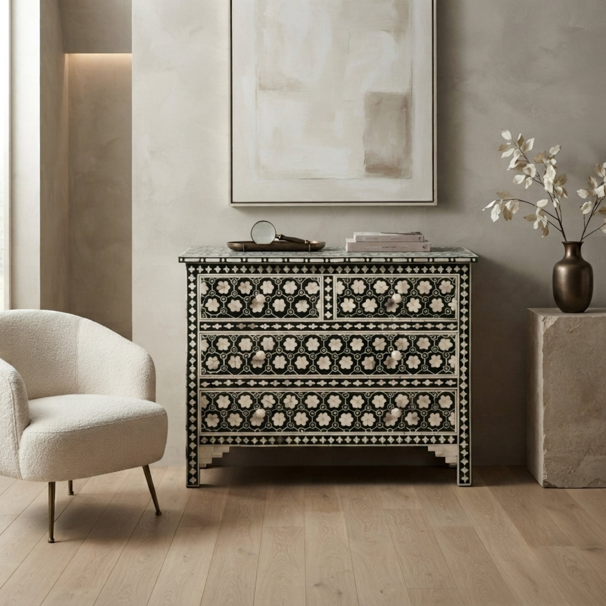 A black and white mother-of-pearl inlaid chest of drawers styled in a modern living room setting with a beige armchair, a concrete side table, and a framed abstract print on the wall.
