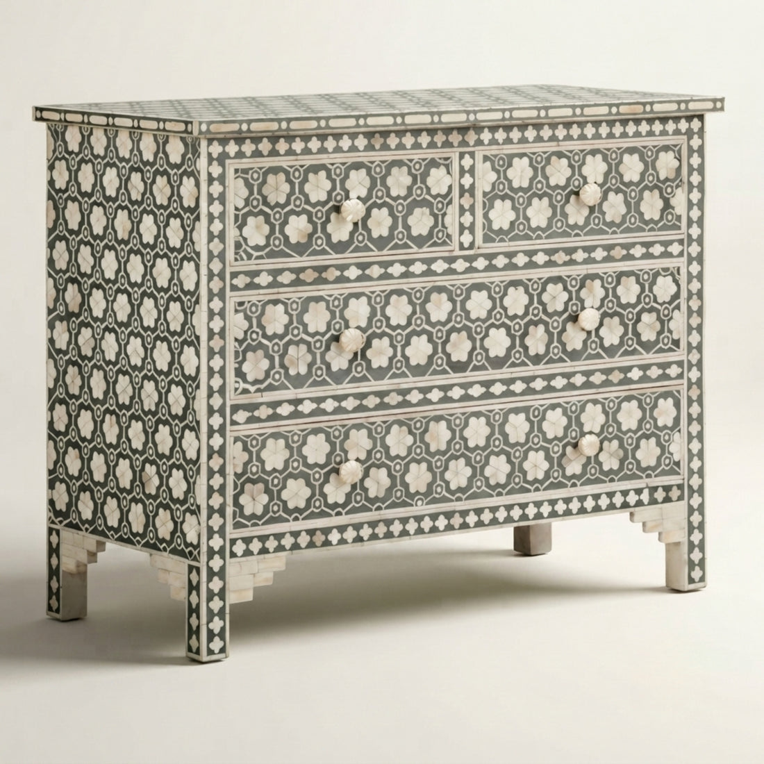 A decorative chest of drawers with an intricate black and white floral bone-inlay pattern, styled in a warm beige room next to a gray armchair with a vase of dried branches on top.