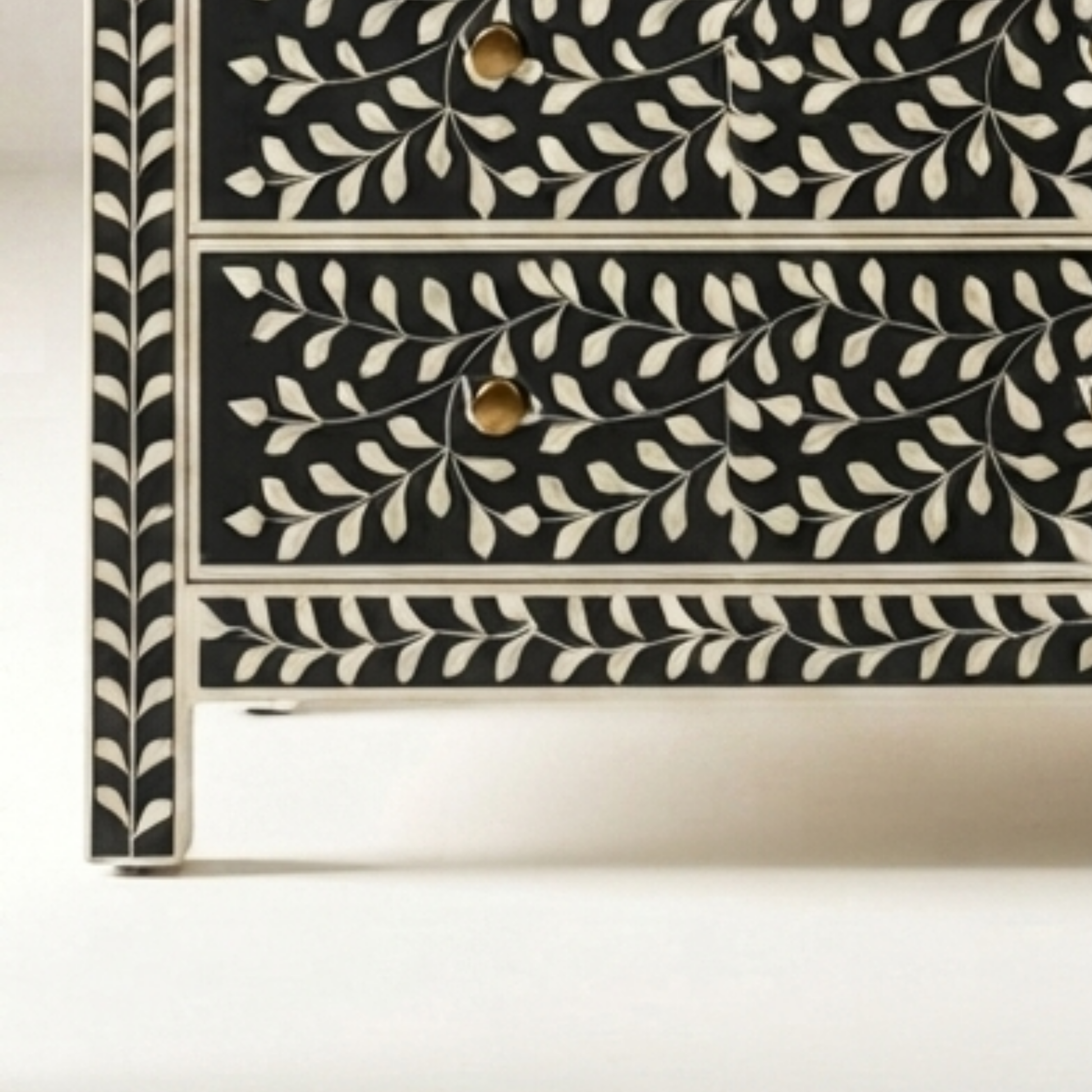Close-up of a black and cream bone-inlay chest of drawers showing two drawers with gold-toned round knobs and a detailed leaf pattern.