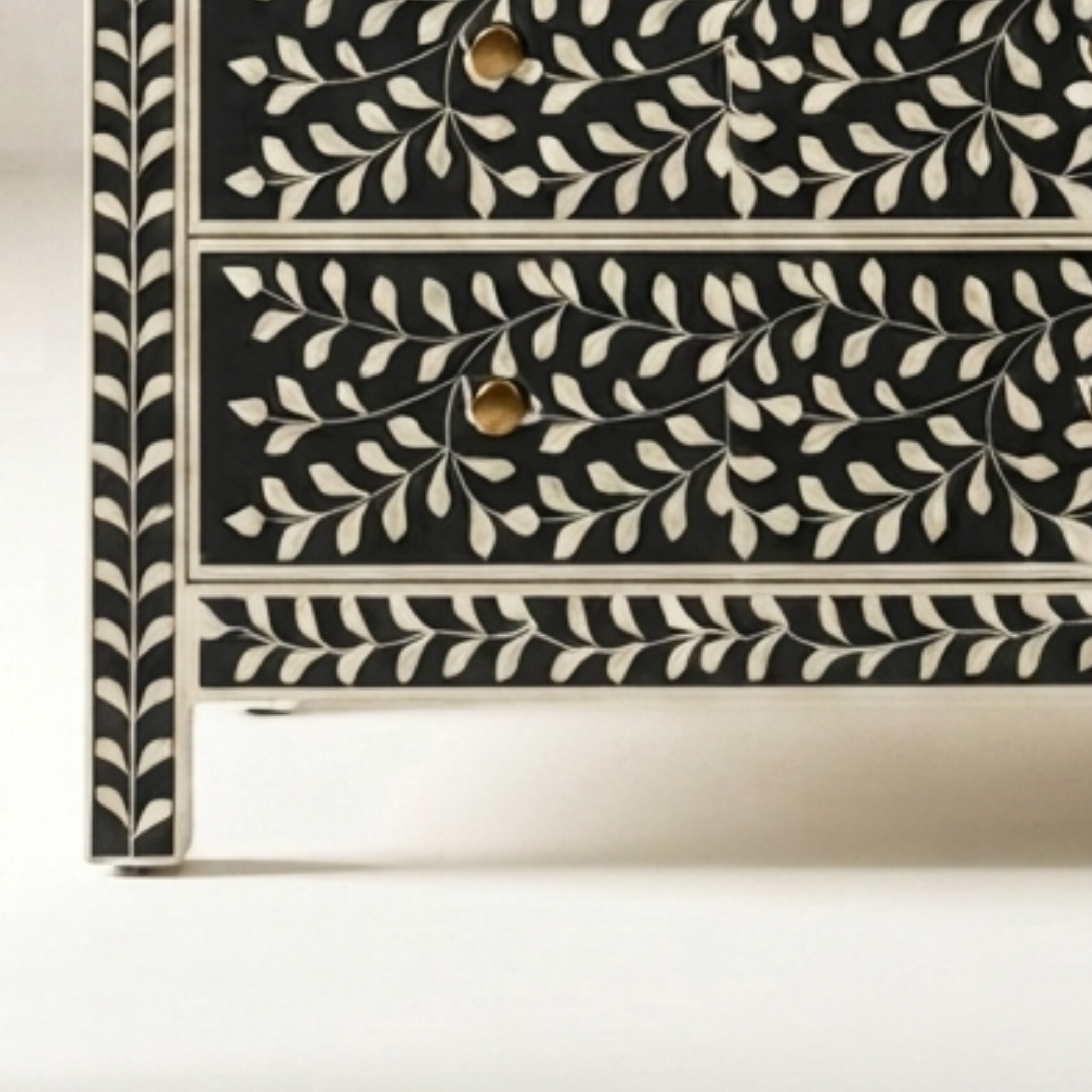 Close-up of a black and cream bone-inlay chest of drawers showing two drawers with gold-toned round knobs and a detailed leaf pattern.