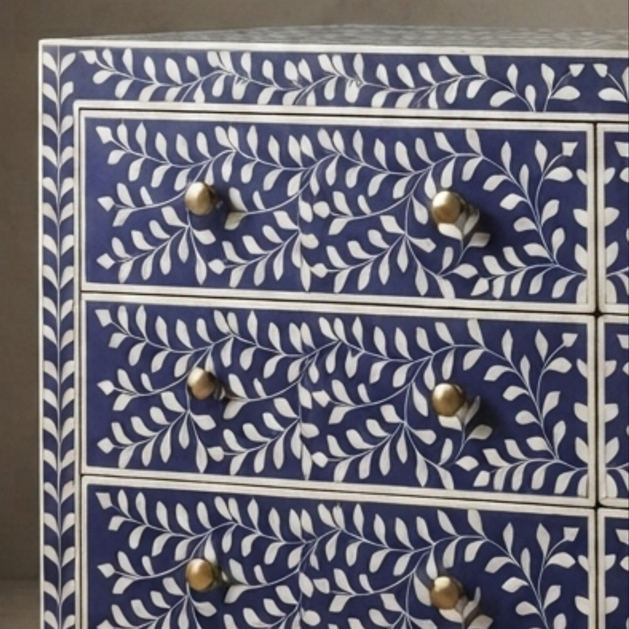 A close-up of a blue and white chest of drawers with an intricate leaf and vine pattern on the front, featuring three visible drawers with round metallic knobs.