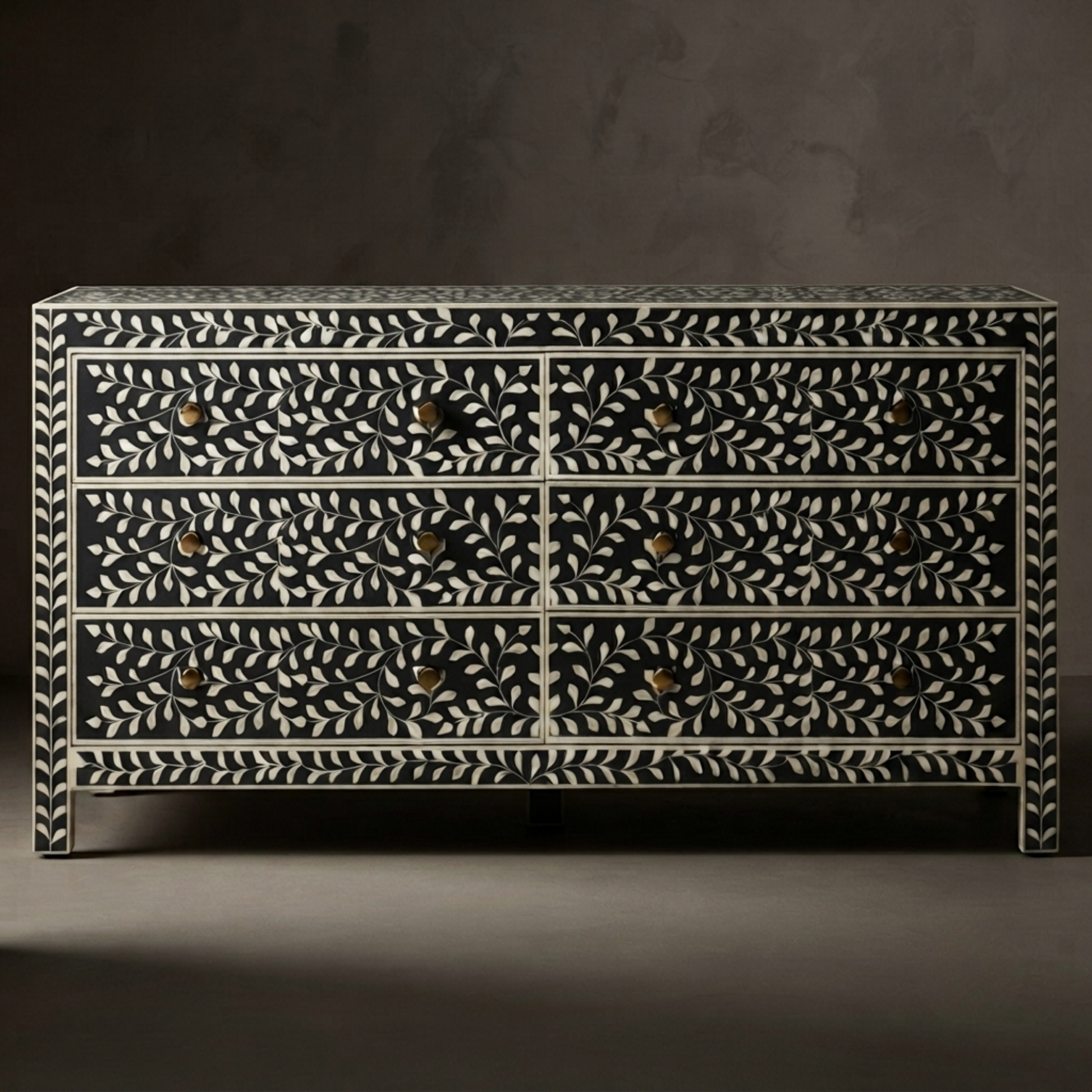A six-drawer dresser with an intricate black and white bone-inlay leaf pattern and small brass knobs, set against a dark gray background.