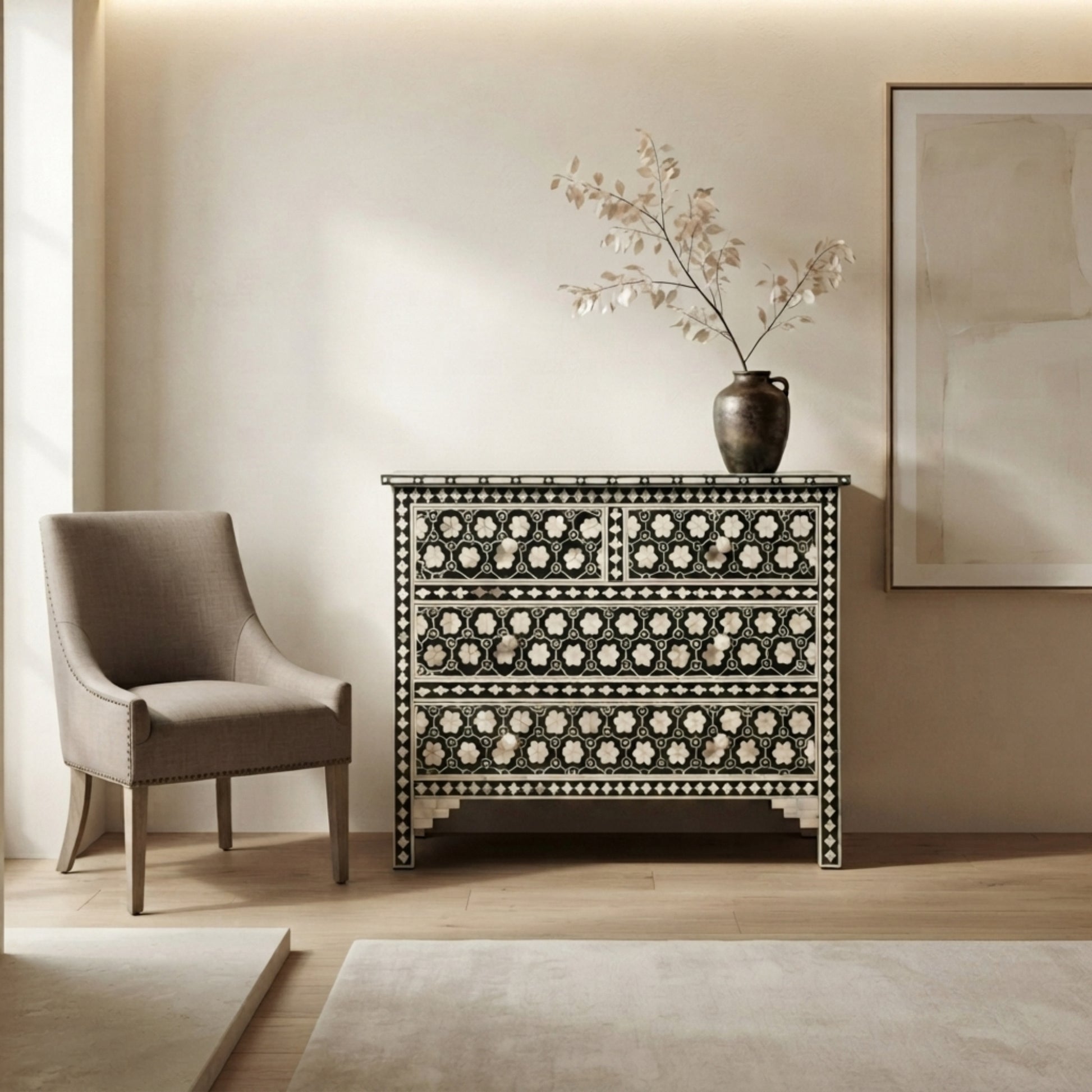A black and white mother-of-pearl inlaid chest of drawers stands in a styled room next to a beige armchair, with a dark ceramic vase holding dried branches on top.