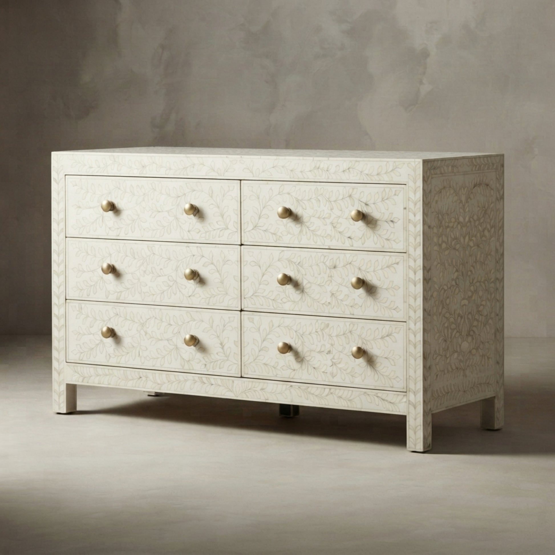 A six-drawer dresser with a textured, off-white bone or shell-embossed finish and small round brass knobs, set against a neutral gray textured studio background.