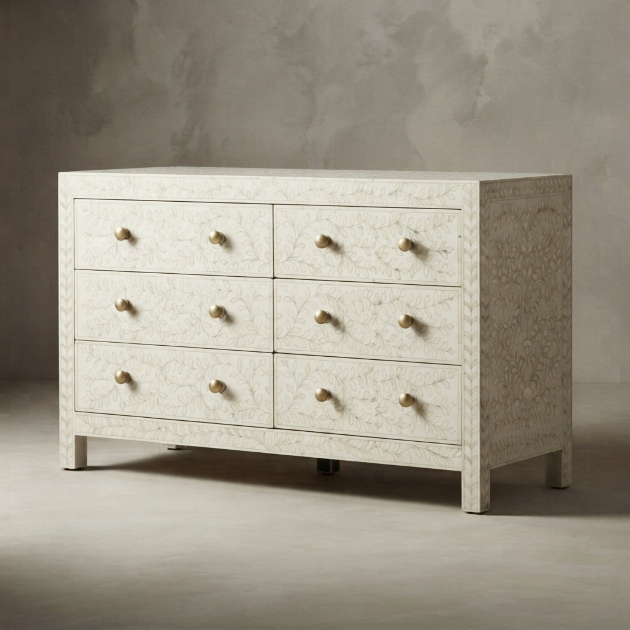 A six-drawer dresser with a textured, off-white bone or shell-embossed finish and small round brass knobs, set against a neutral gray textured studio background.