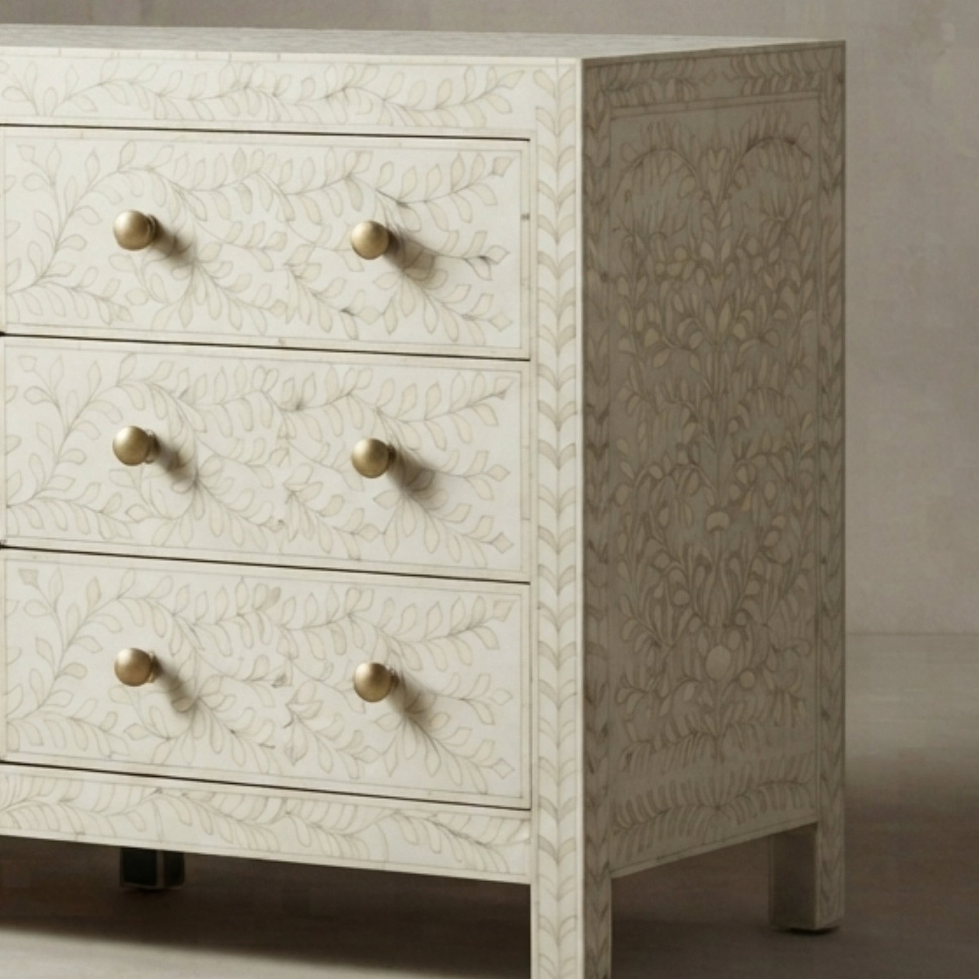 An off-white bone-in-wood chest of drawers with three drawers, featuring a delicate floral inlay pattern and round brass knobs, shown against a neutral background.