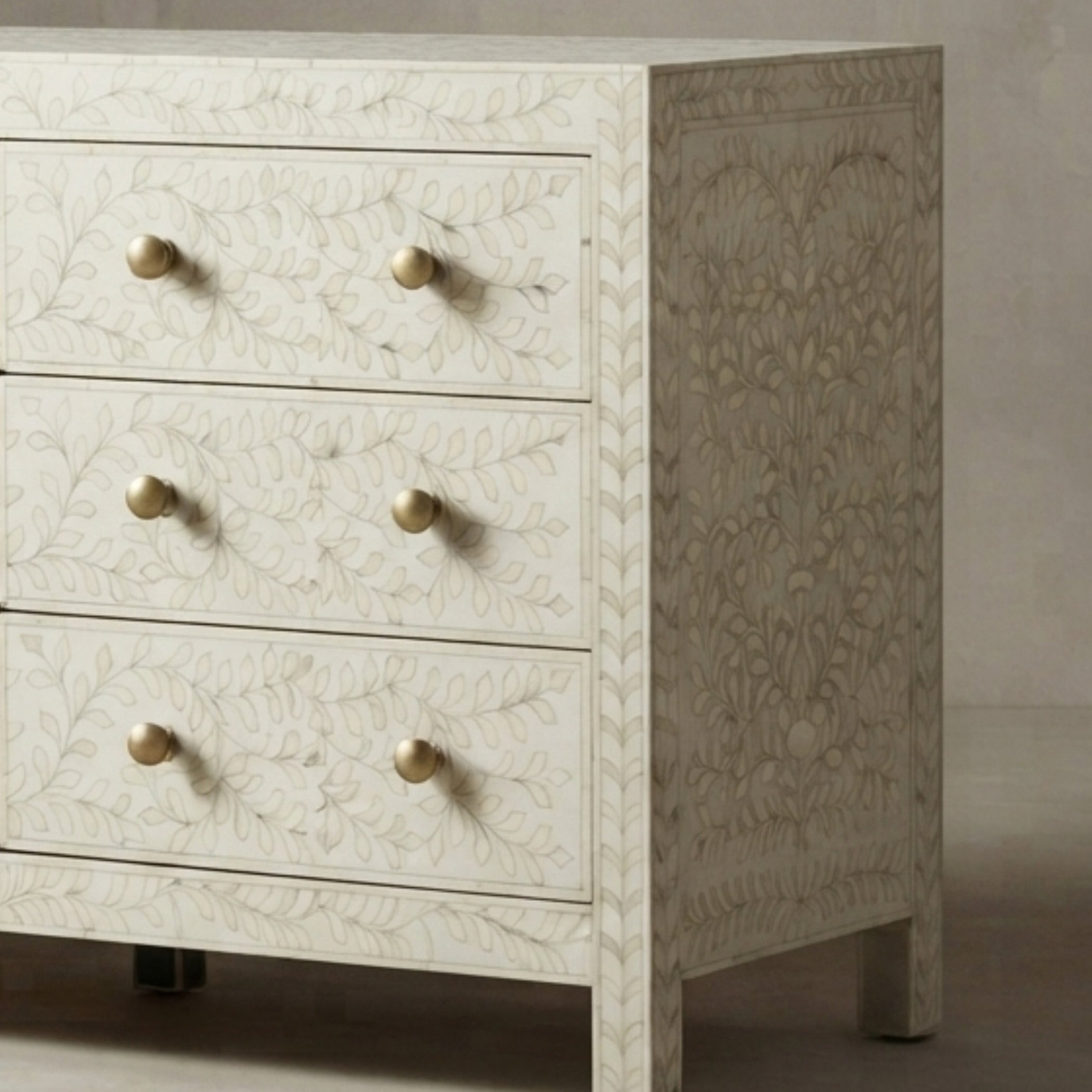 An off-white bone-in-wood chest of drawers with three drawers, featuring a delicate floral inlay pattern and round brass knobs, shown against a neutral background.