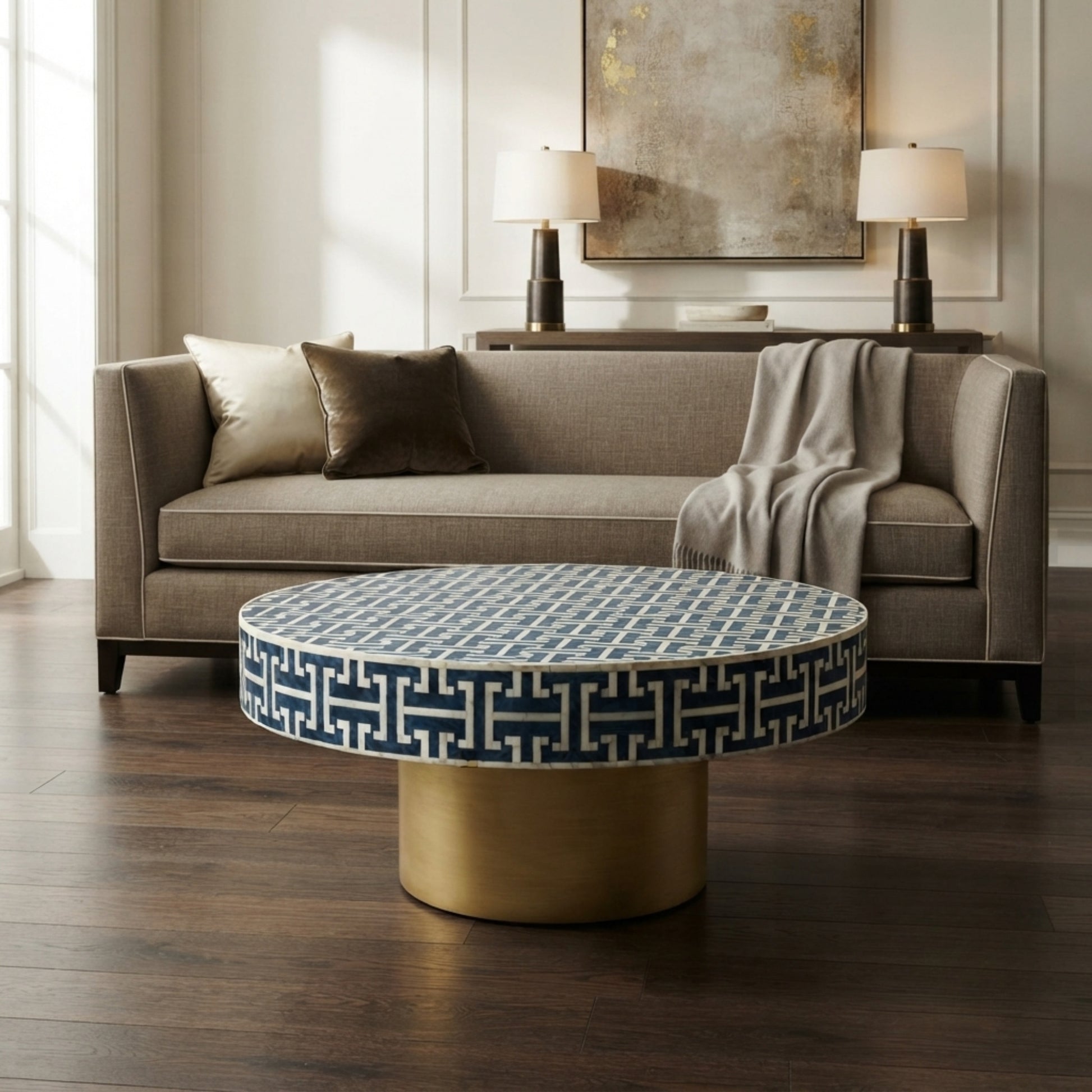 A round coffee table with a navy blue and white geometric patterned top and a cylindrical gold base, positioned in front of a gray fabric sofa in a modern living room setting.