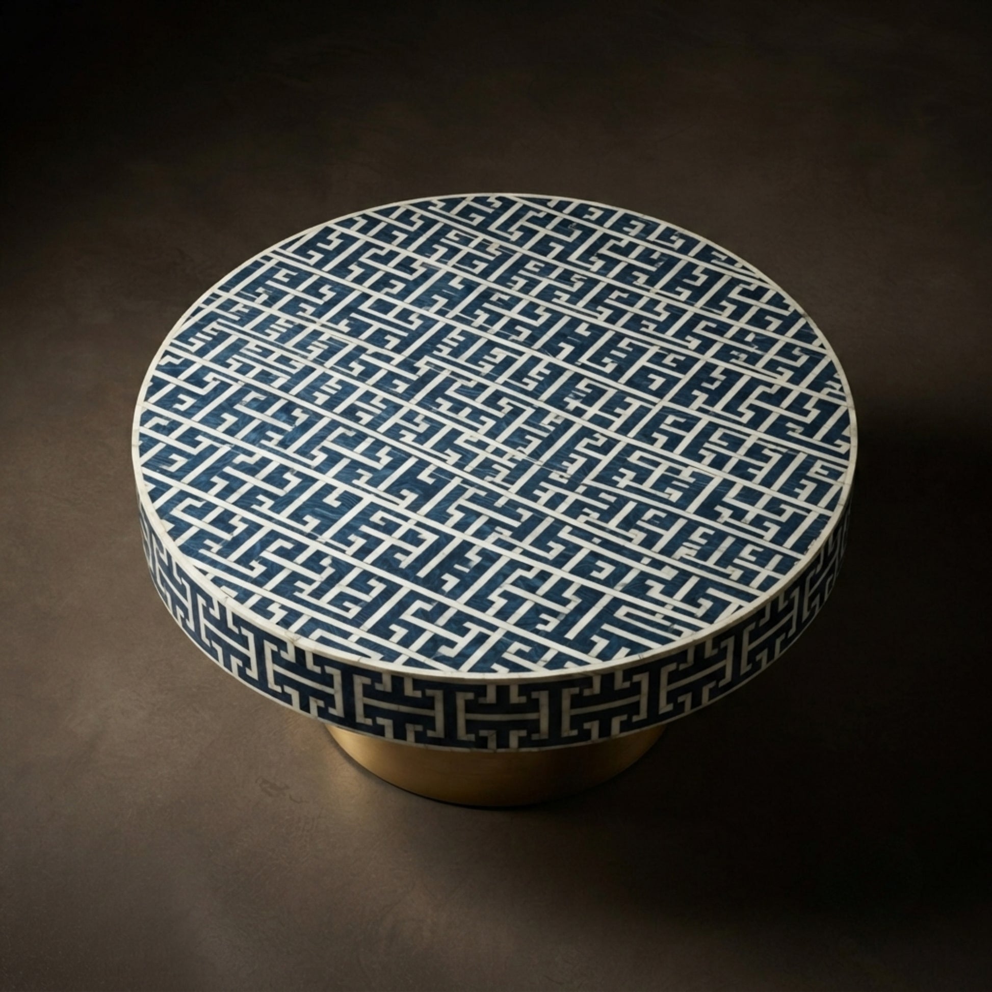 A round low coffee table with a dark blue and white geometric Greek key pattern on the top and a matching patterned rim, supported by a gold-finished cylindrical base, set against a dark brown background.