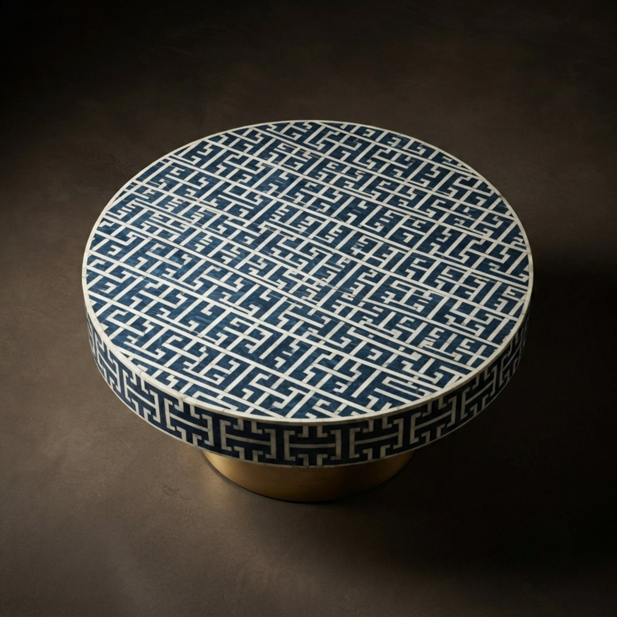 A round low coffee table with a dark blue and white geometric Greek key pattern on the top and a matching patterned rim, supported by a gold-finished cylindrical base, set against a dark brown background.