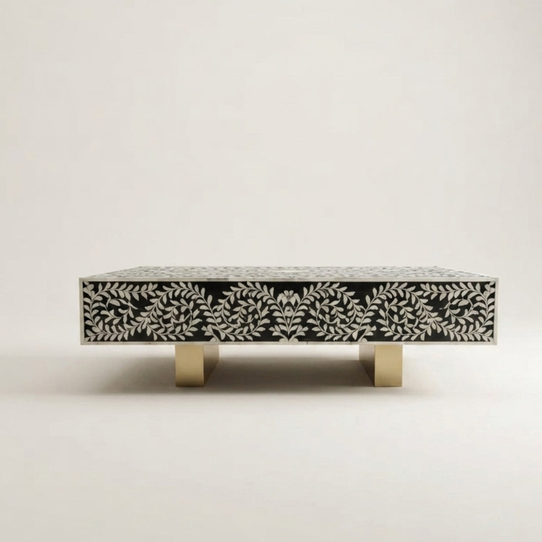 A square modern coffee table with a black and white bone-in-shell leaf pattern and gold legs, styled in a bright, contemporary living room next to a cream bouclé sofa.