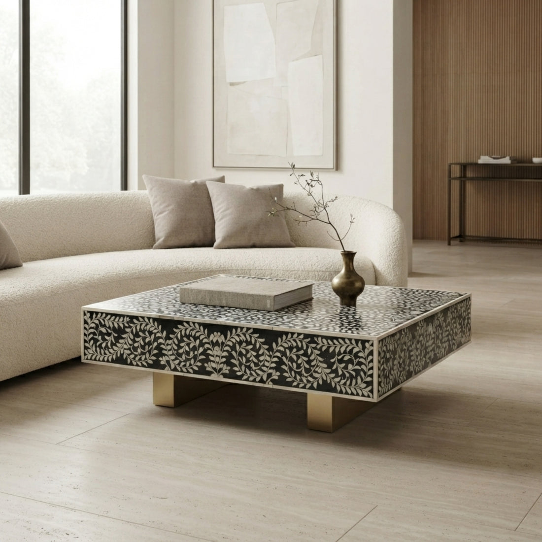 A square modern coffee table with a black and white bone-in-shell leaf pattern and gold legs, styled in a bright, contemporary living room next to a cream bouclé sofa.