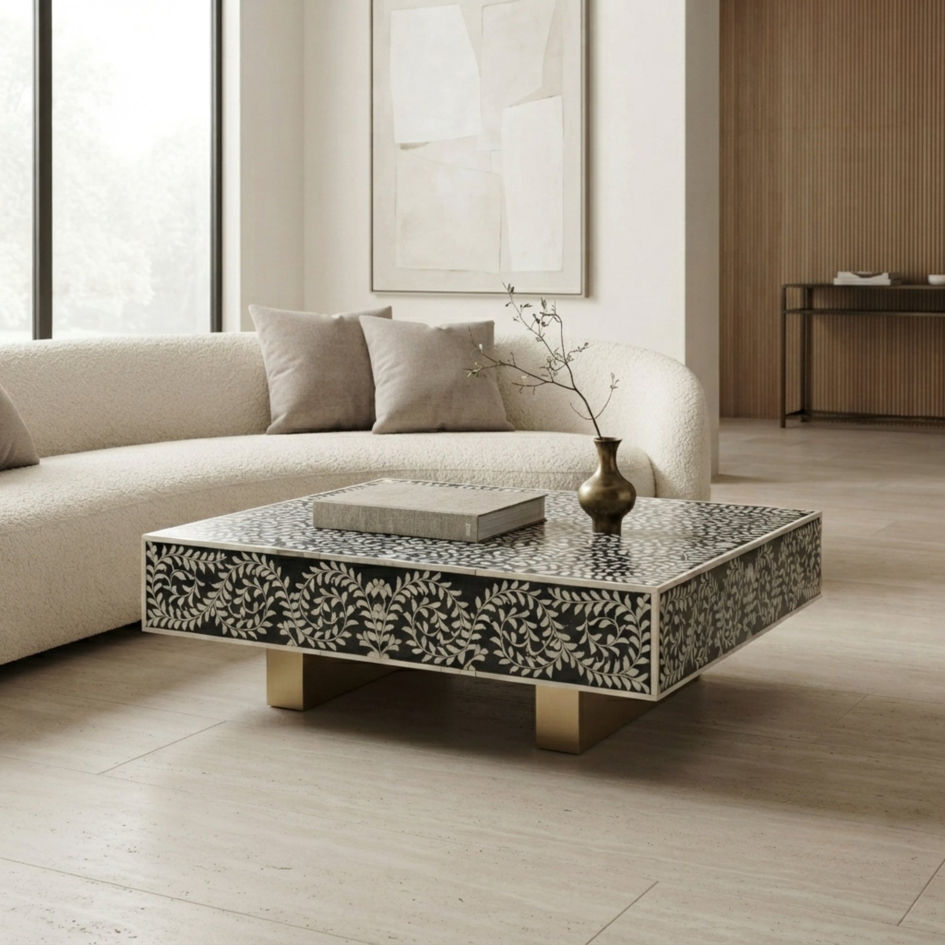 A square modern coffee table with a black and white bone-in-shell leaf pattern and gold legs, styled in a bright, contemporary living room next to a cream bouclé sofa.