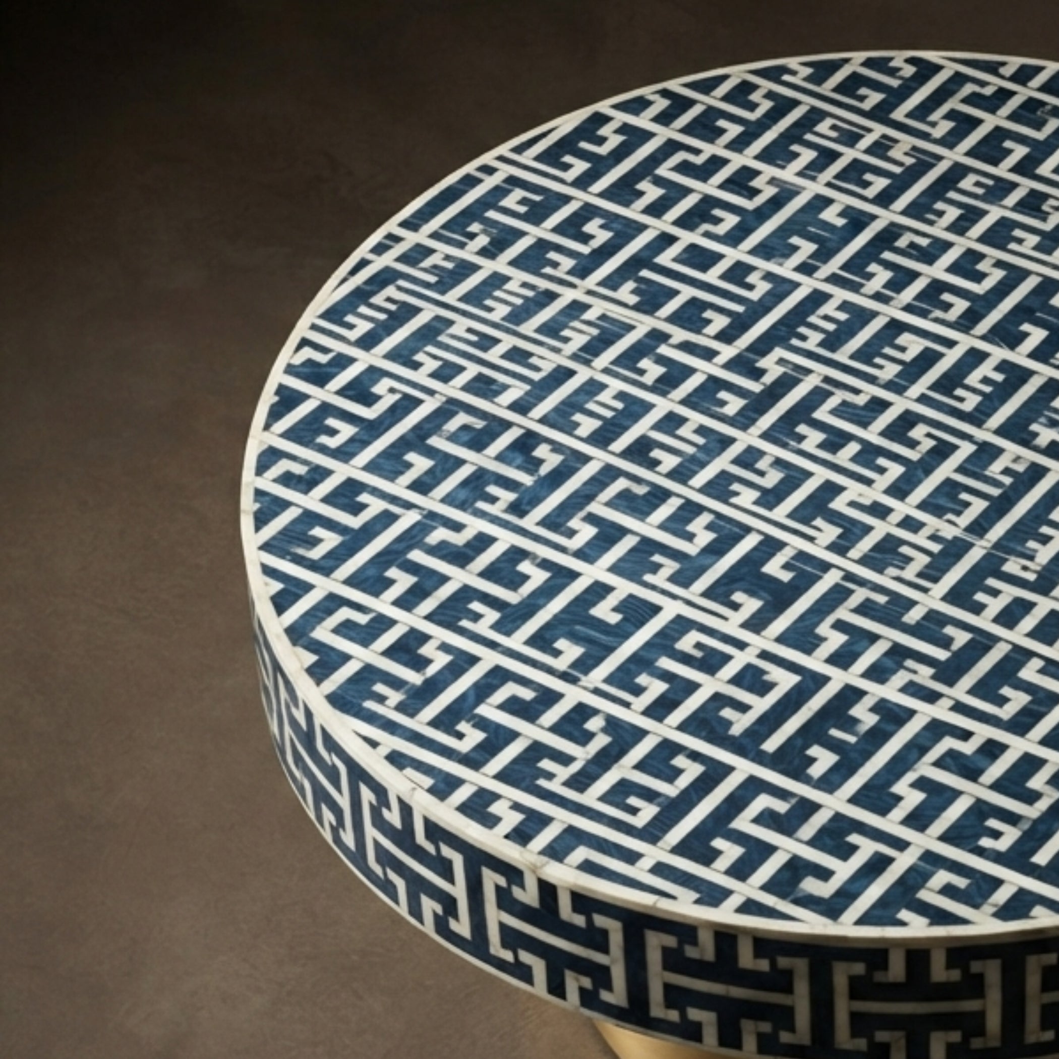 A close-up of a round table with a dark blue and white geometric Greek key pattern on the tabletop and side, set against a dark brown textured background.