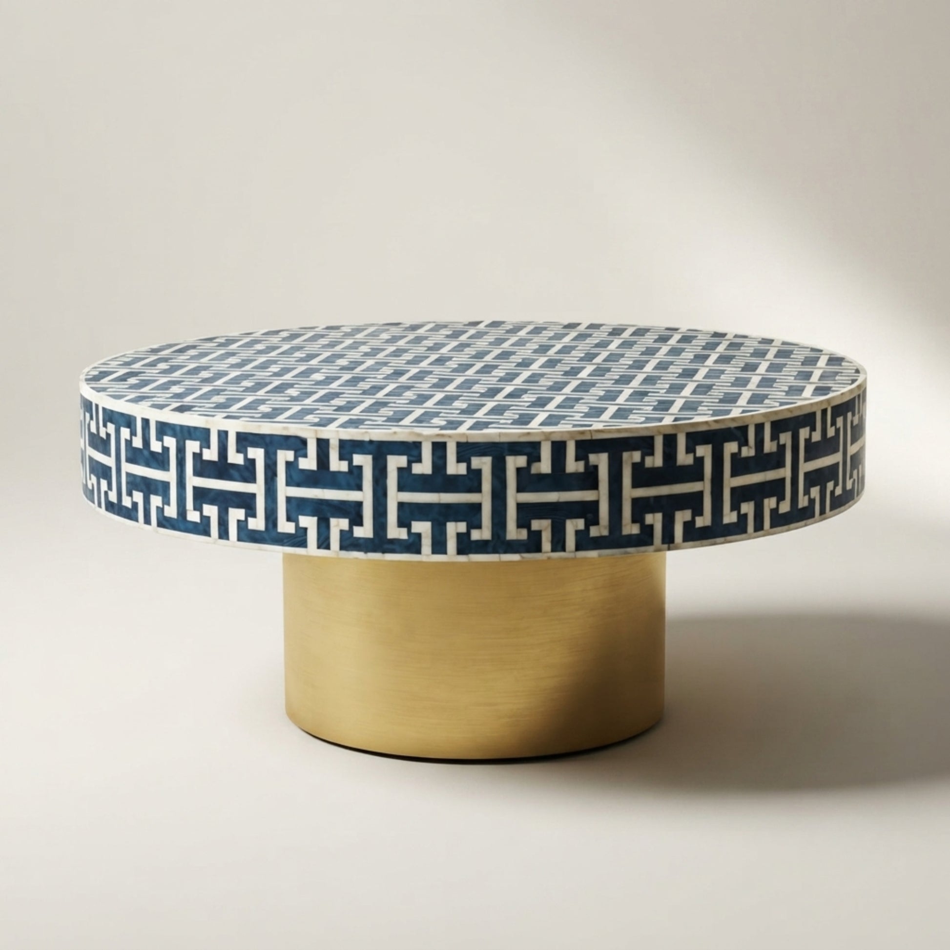 A round coffee table with a blue and white geometric patterned top and a brushed gold cylindrical base, photographed on a neutral light gray studio background.