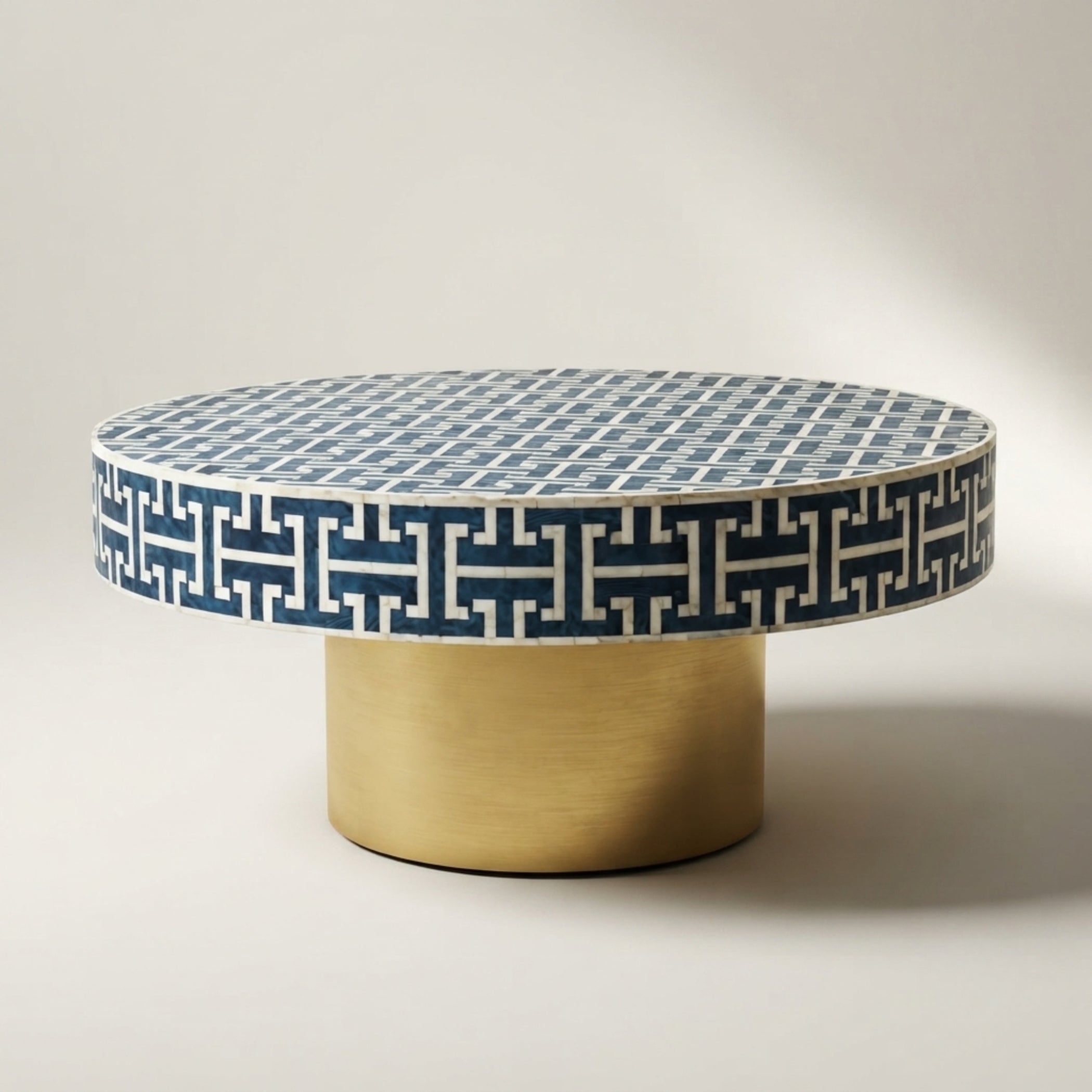 A round coffee table with a blue and white geometric patterned top and a brushed gold cylindrical base, photographed on a neutral light gray studio background.