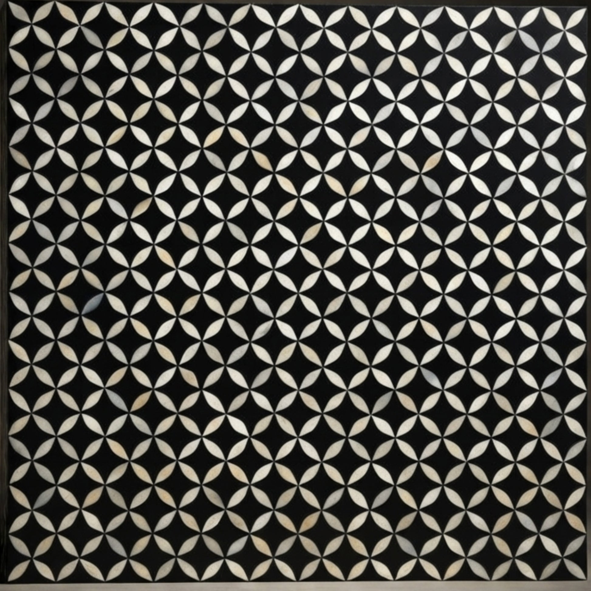 A square black panel featuring a repeating geometric pattern of interlocking white and beige petal-like shapes, bordered by a thin dark frame.