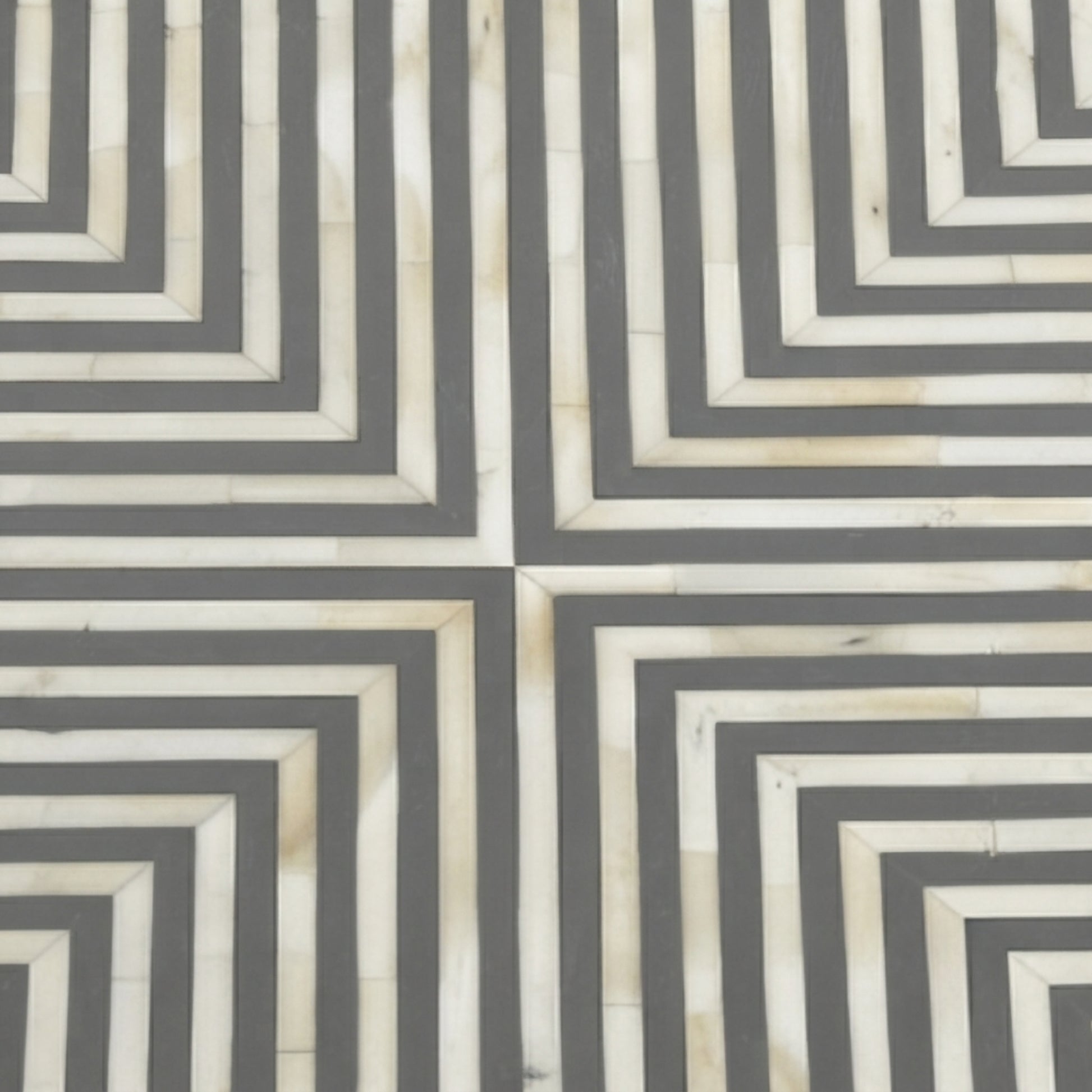 A close-up of a geometric patterned surface with alternating dark gray and warm off-white concentric rectangular stripes, forming a repeating maze-like geometric design.