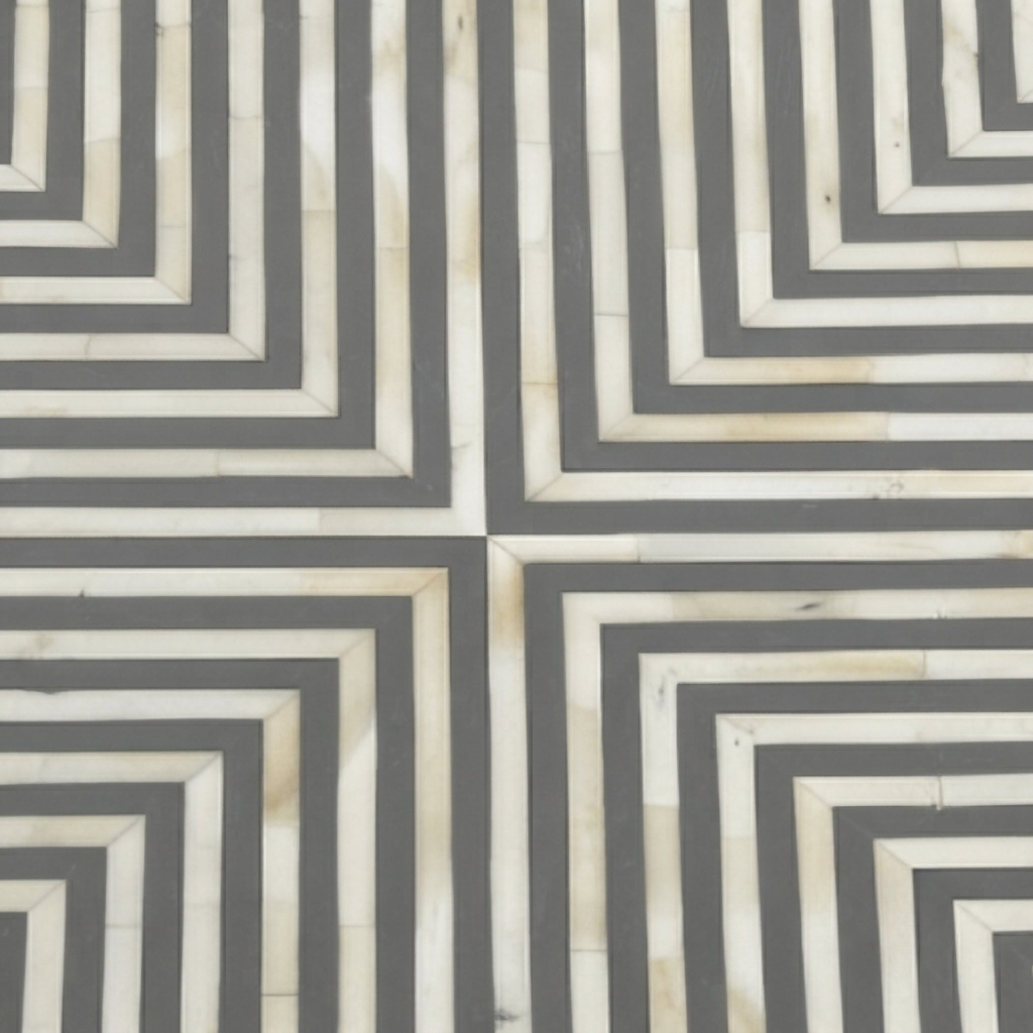 A close-up of a geometric patterned surface with alternating dark gray and warm off-white concentric rectangular stripes, forming a repeating maze-like geometric design.