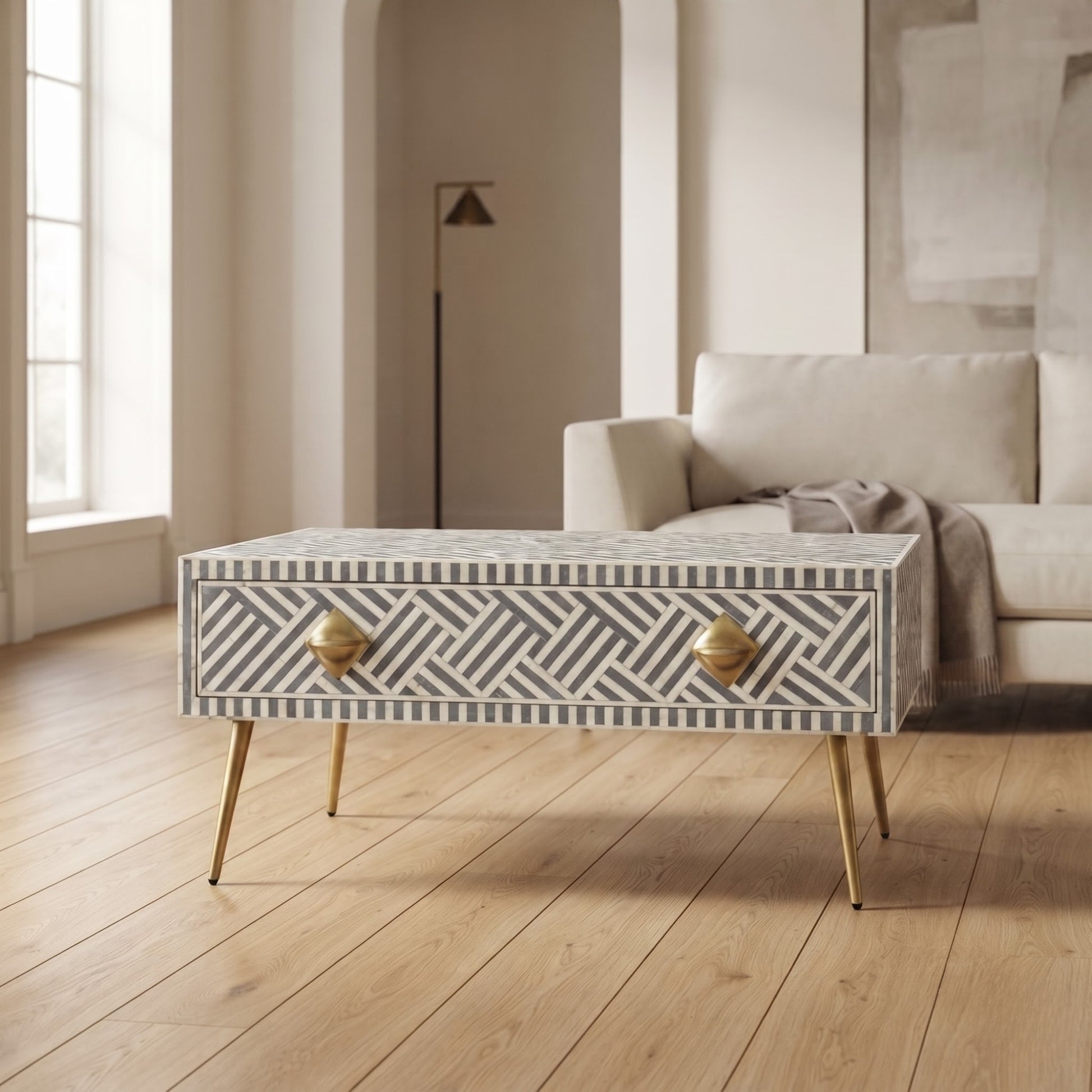A rectangular coffee table with a geometric bone-inlay pattern in gray and white, featuring two brass drawer pulls and slim gold metal legs, set in a bright, modern living room.