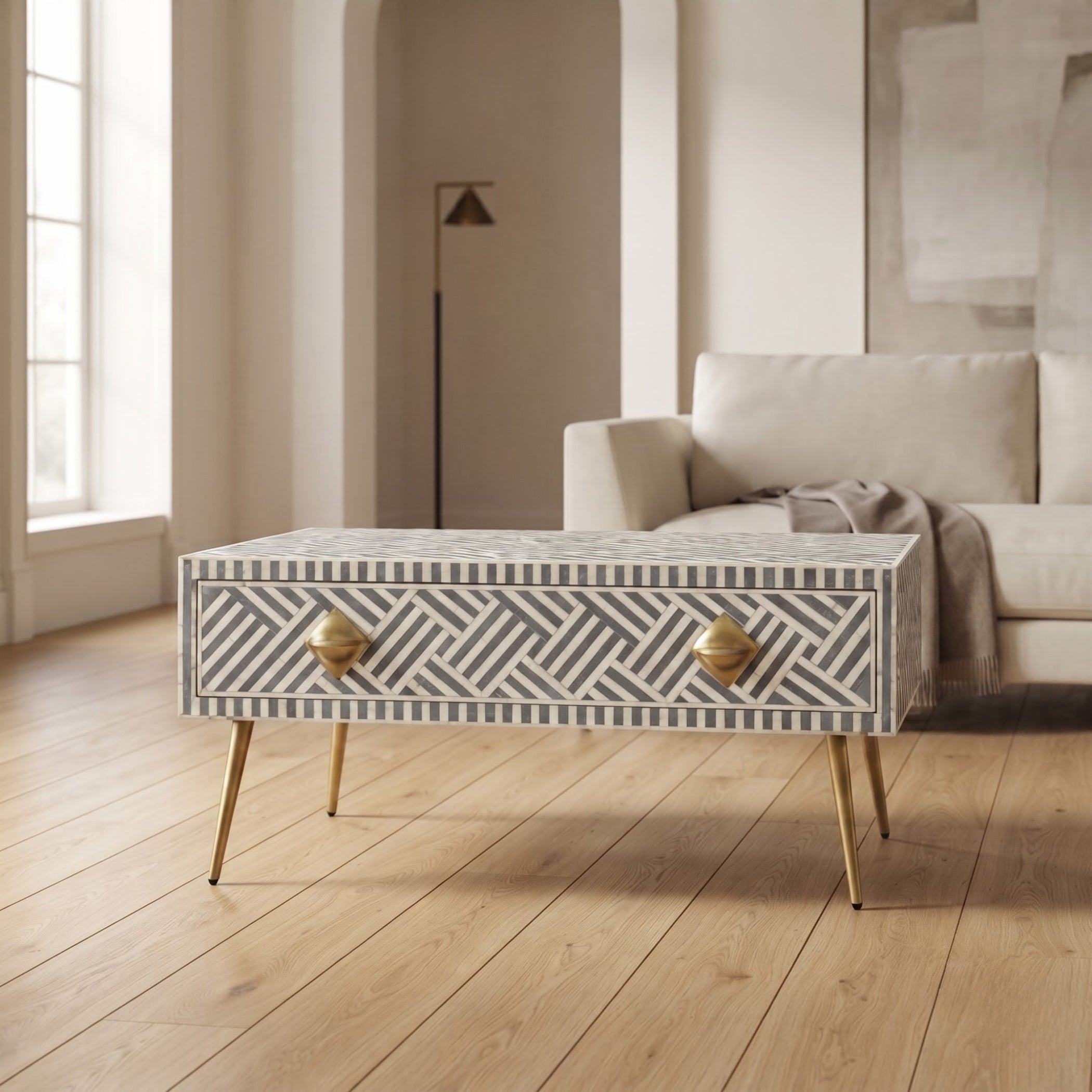 A rectangular coffee table with a geometric bone-inlay pattern in gray and white, featuring two brass drawer pulls and slim gold metal legs, set in a bright, modern living room.
