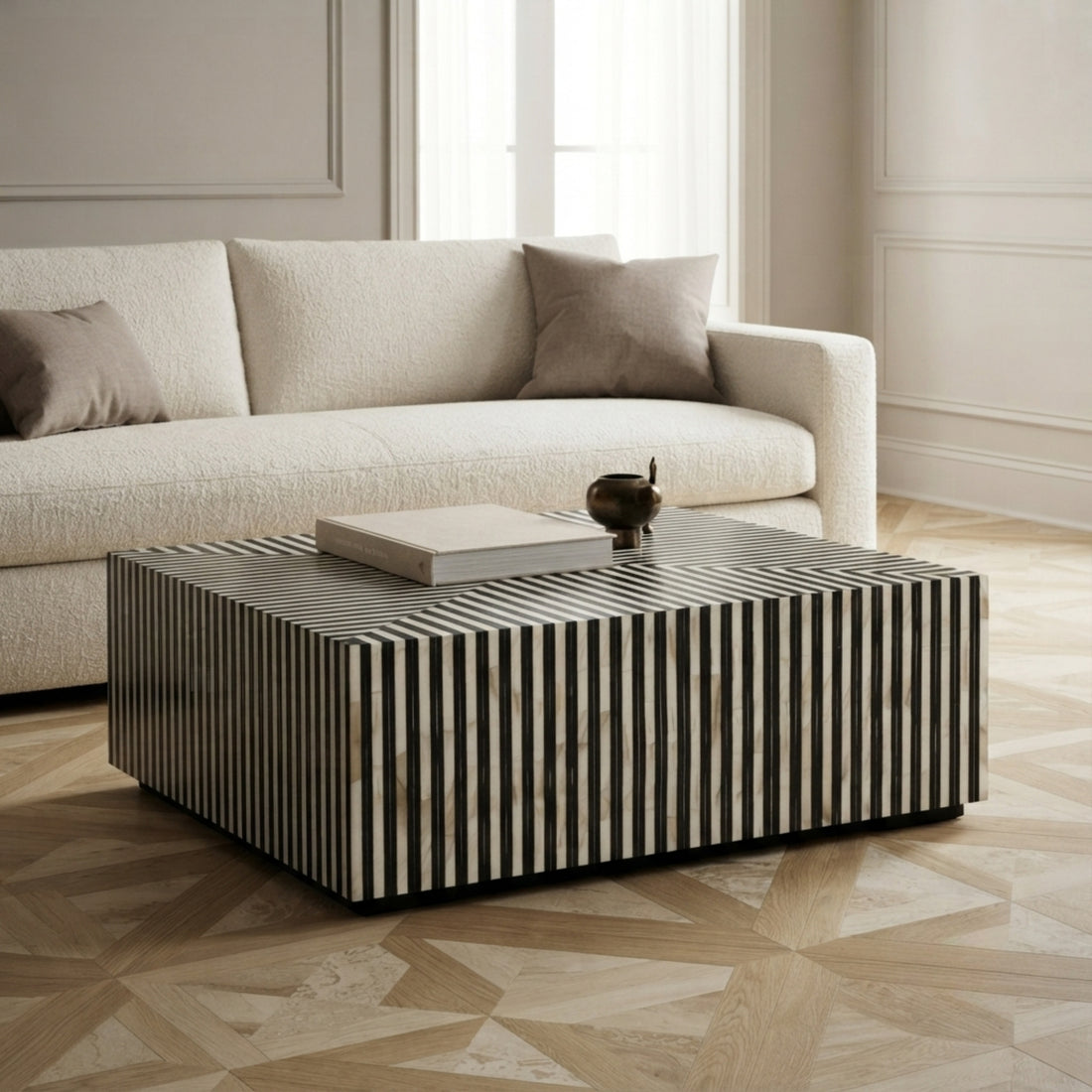 A modern square coffee table with vertical black and white striped inlay, styled in a bright living room next to a cream-colored sofa.