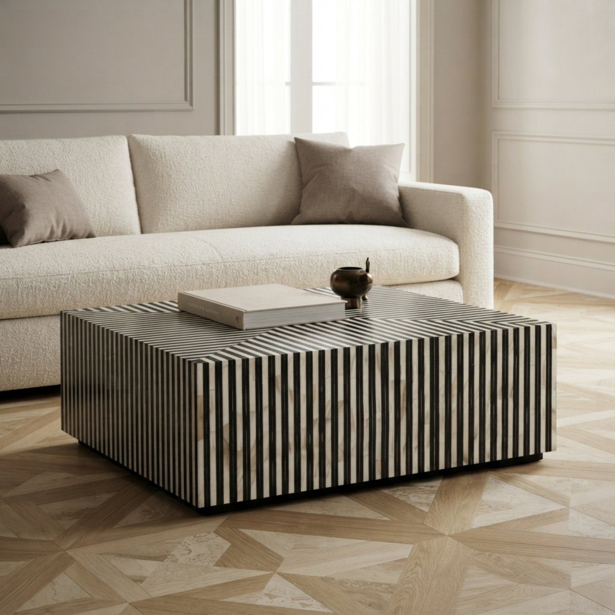 A modern square coffee table with vertical black and white striped inlay, styled in a bright living room next to a cream-colored sofa.