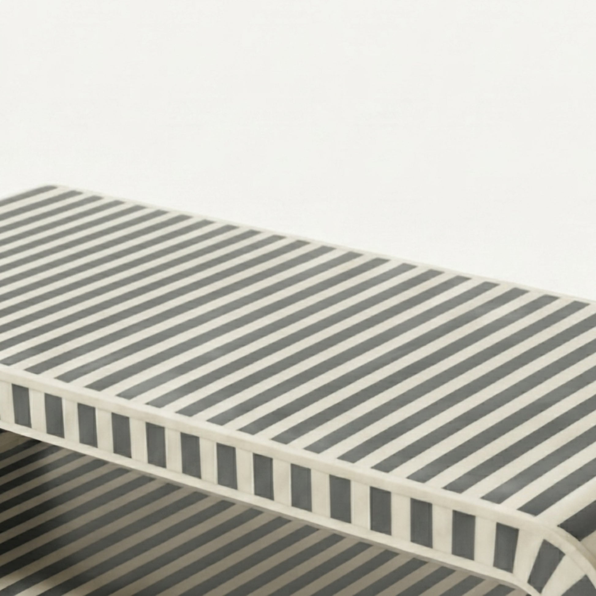 A close-up of a rectangular table or bench with a striped pattern in dark gray and off-white, showing the side profile and edge details against a plain light background.