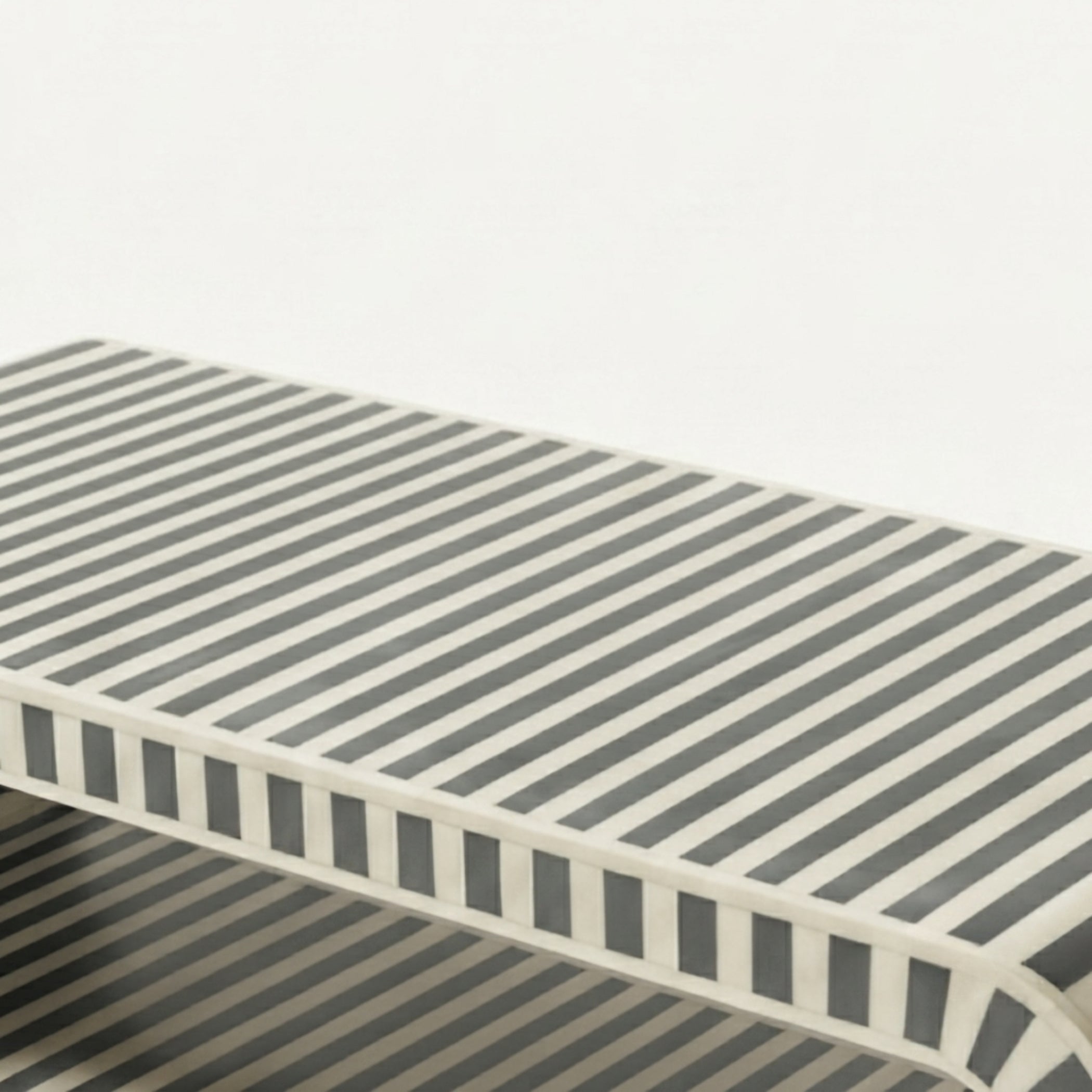 A close-up of a rectangular table or bench with a striped pattern in dark gray and off-white, showing the side profile and edge details against a plain light background.