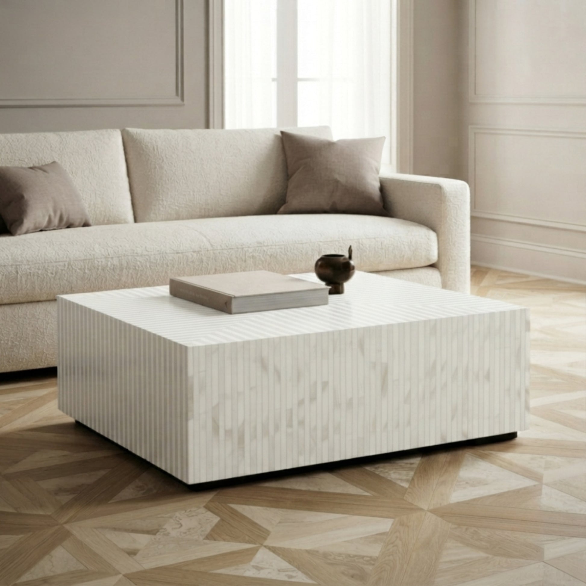 A modern square coffee table with a textured white and light beige wood-grain finish, styled in a living room setting with a cream sofa and geometric wood flooring.