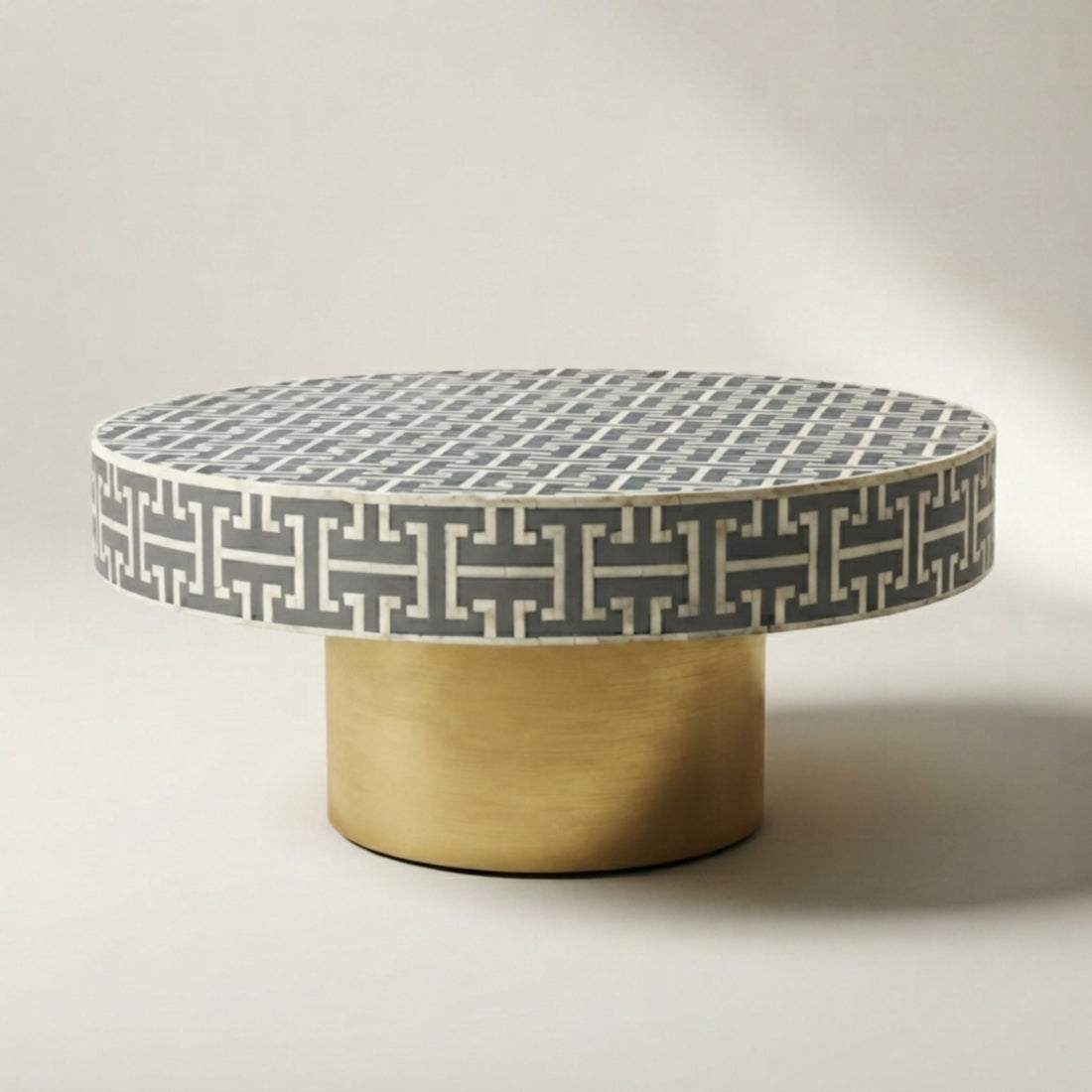 A round coffee table with a gray and white geometric patterned top and a cylindrical brushed gold base, styled in a modern living room setting.