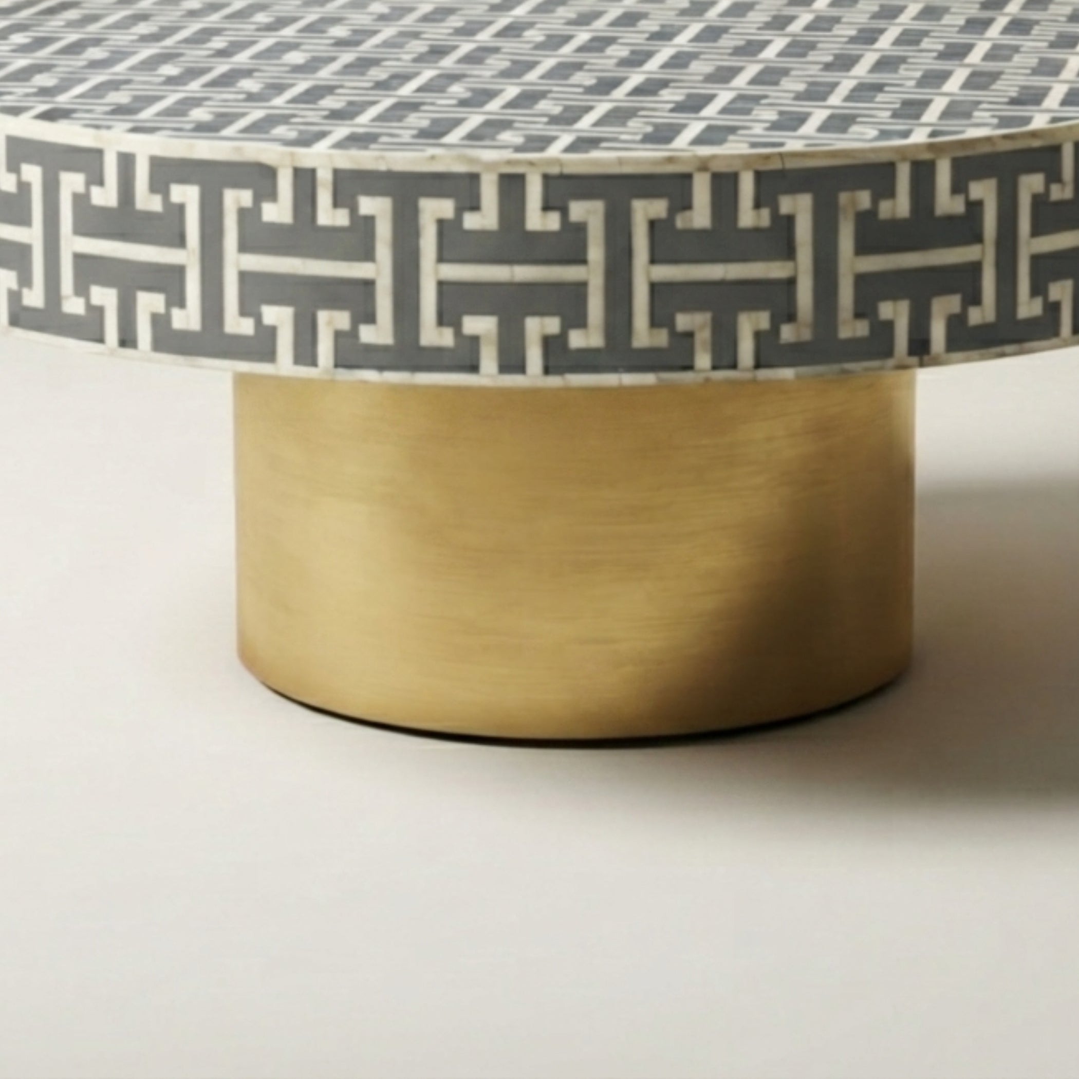 A round accent table featuring a gray and white geometric inlay pattern on the top and a brushed gold cylindrical base, shown on a neutral light gray background.