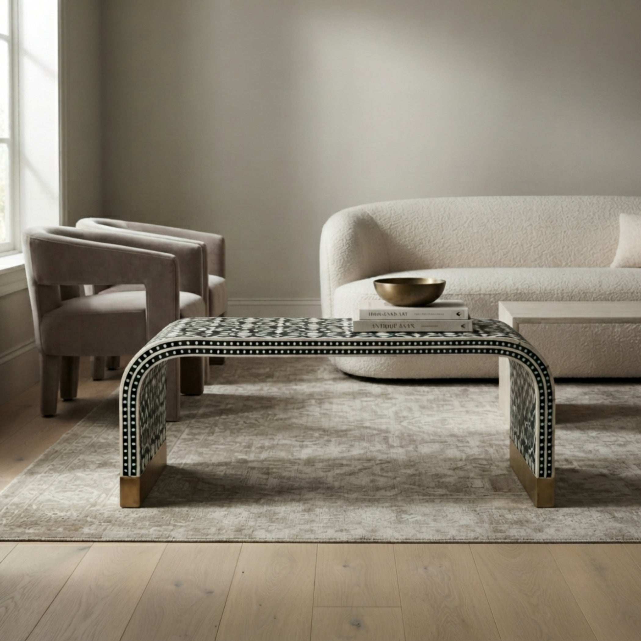 A modern curved coffee table with a black and white mosaic tile pattern and light wood legs, styled in a minimalist living room with a beige sofa, armchair, and textured rug.