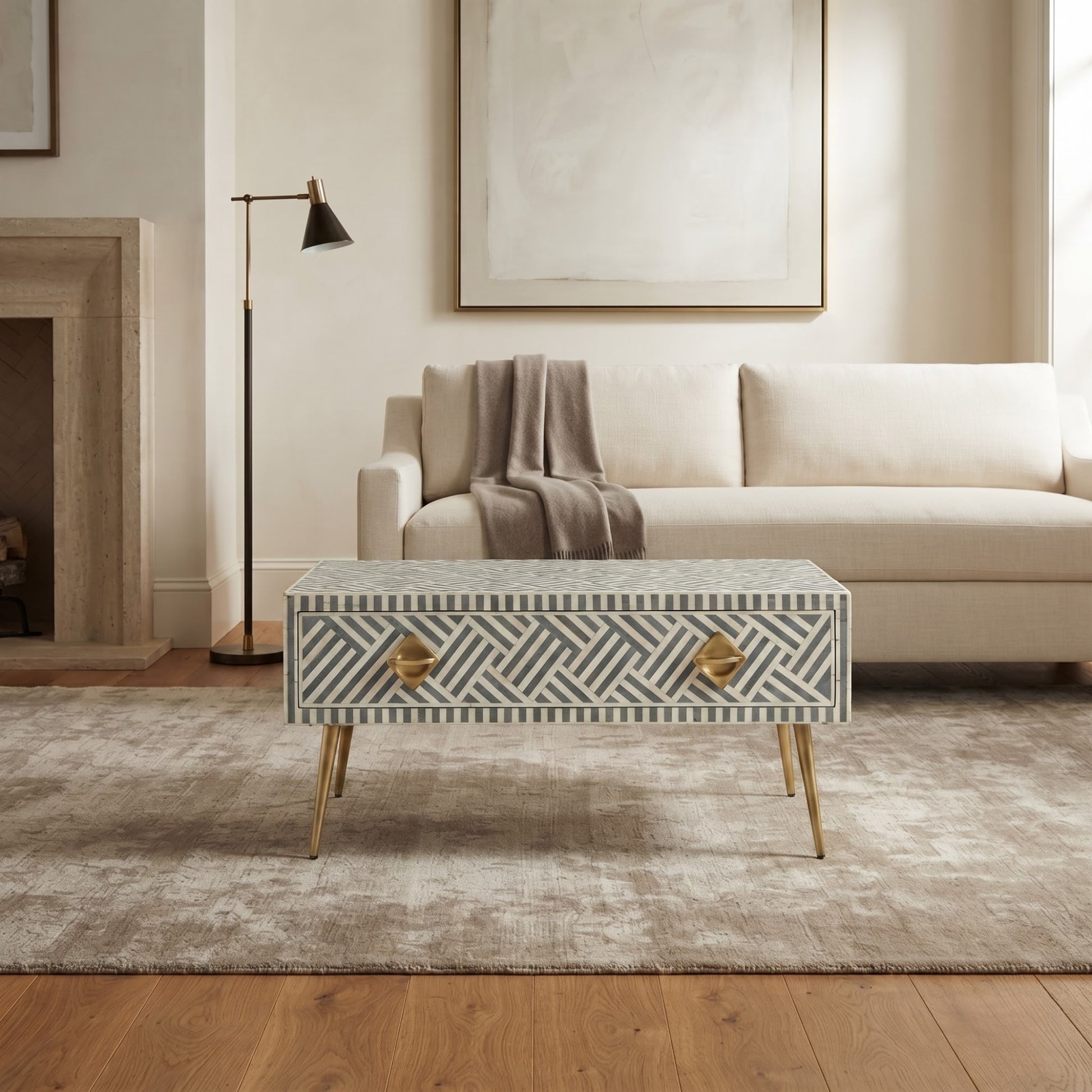 A rectangular coffee table with a geometric bone-inlay pattern in gray and white, featuring two gold-finished drawer pulls and gold tapered legs, set in a modern living room next to a cream sofa.