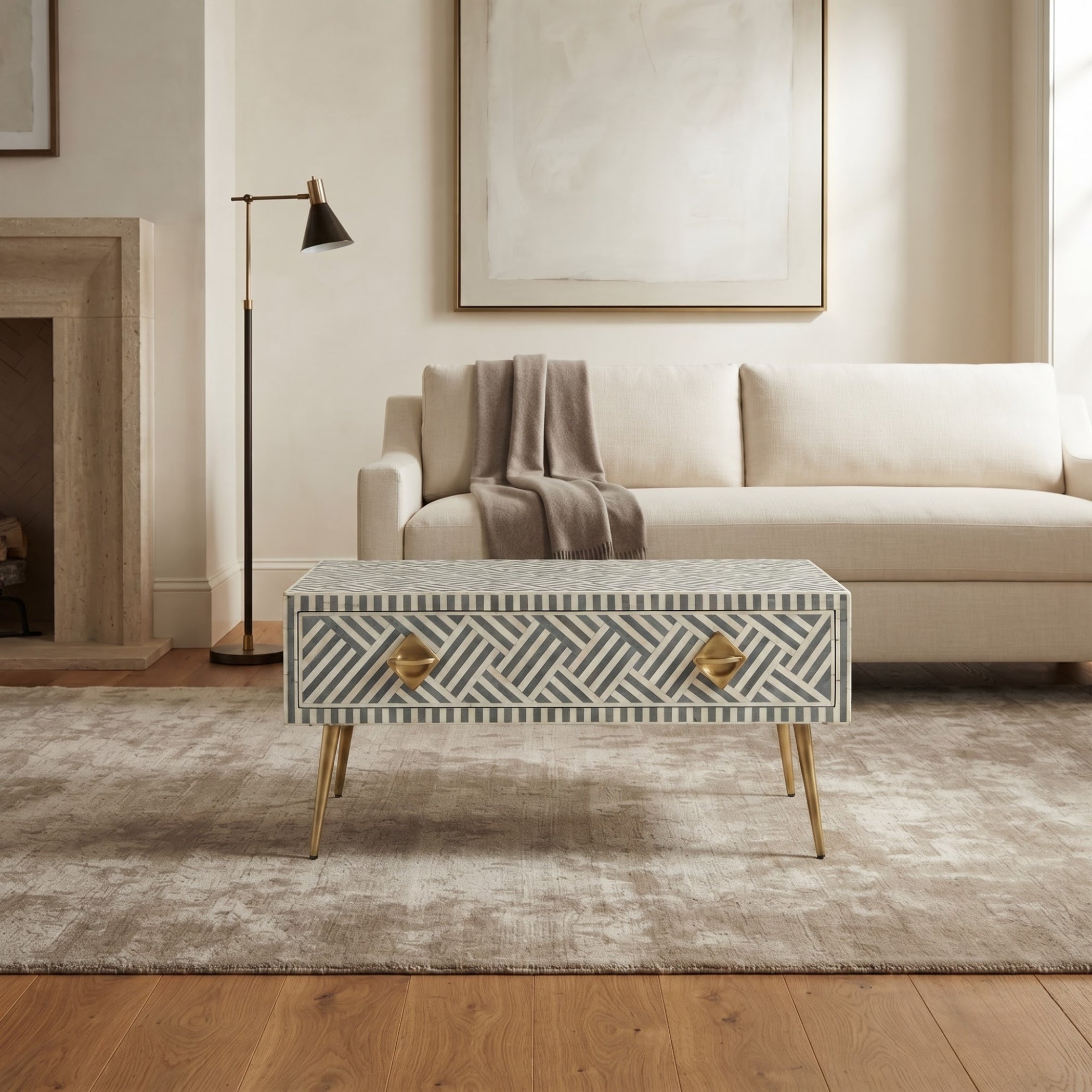A rectangular coffee table with a geometric bone-inlay pattern in gray and white, featuring two gold-finished drawer pulls and gold tapered legs, set in a modern living room next to a cream sofa.