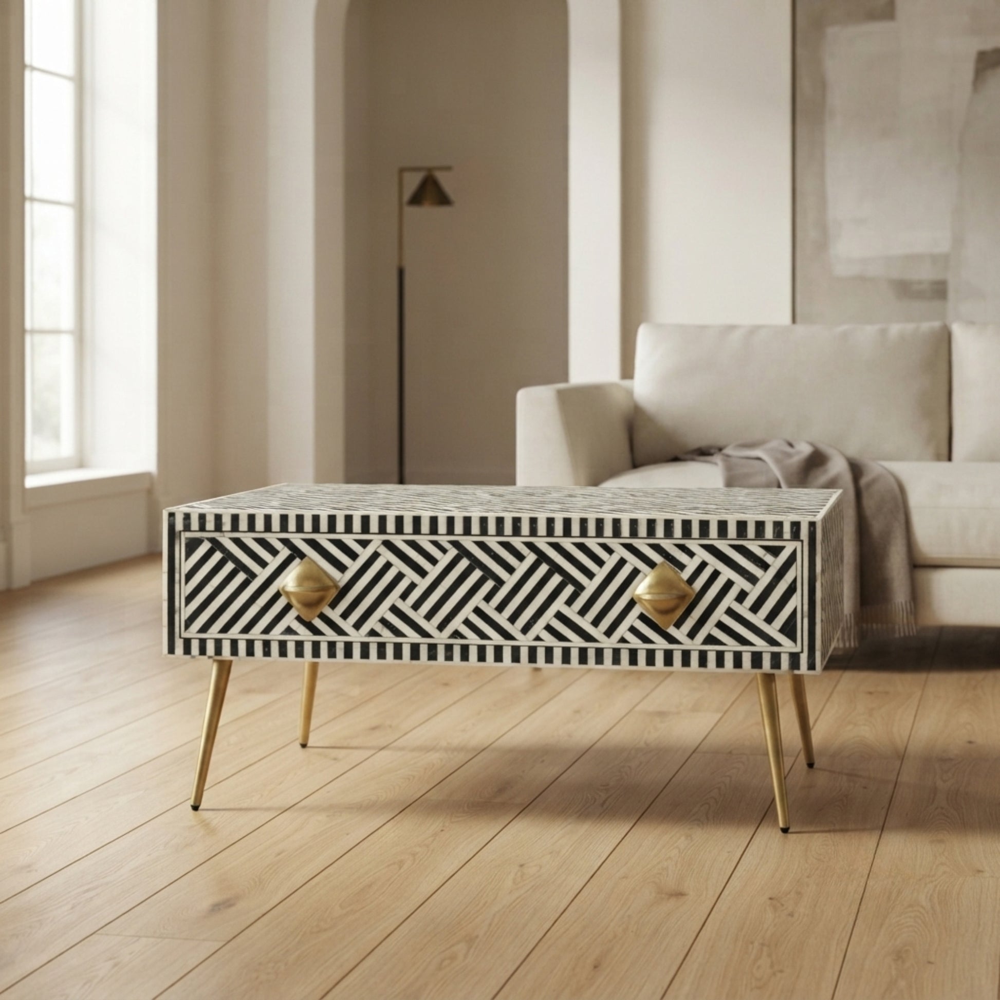A rectangular coffee table with a black and white geometric bone-inlay pattern, gold-finished legs, and round gold drawer pulls, shown in a modern living room setting.