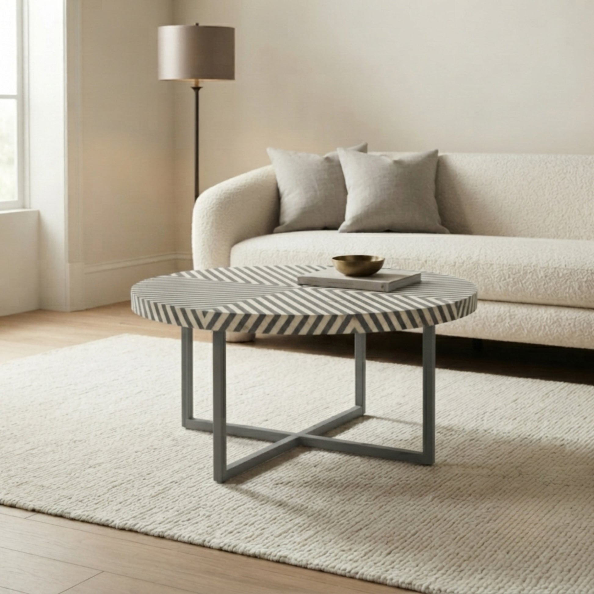 A round coffee table with a black and white striped top and a gray metal frame, styled in a modern living room setting with a cream sofa and textured rug.