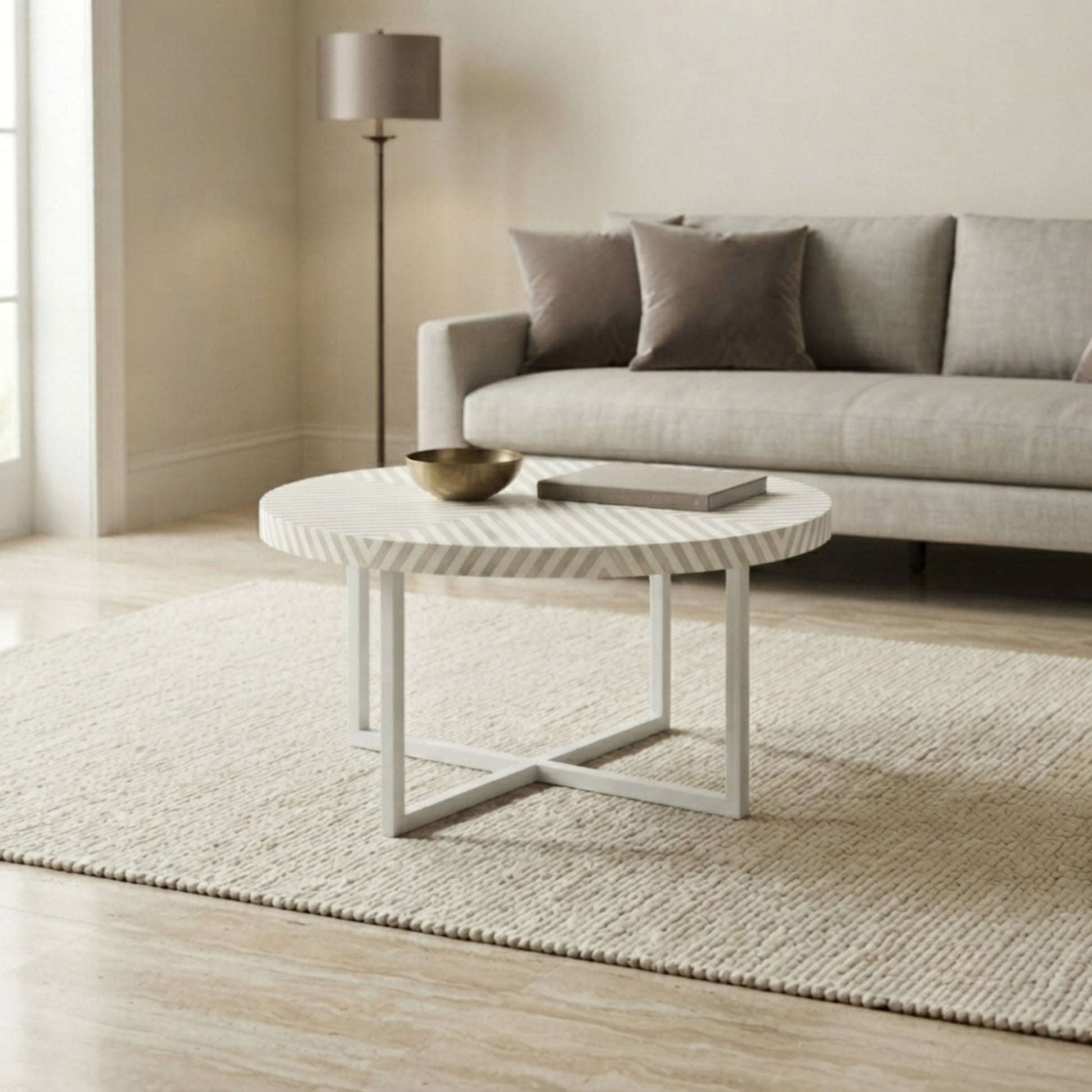 A round white coffee table with a textured top and white metal legs, styled in a modern living room setting on a woven rug in front of a light gray sofa.