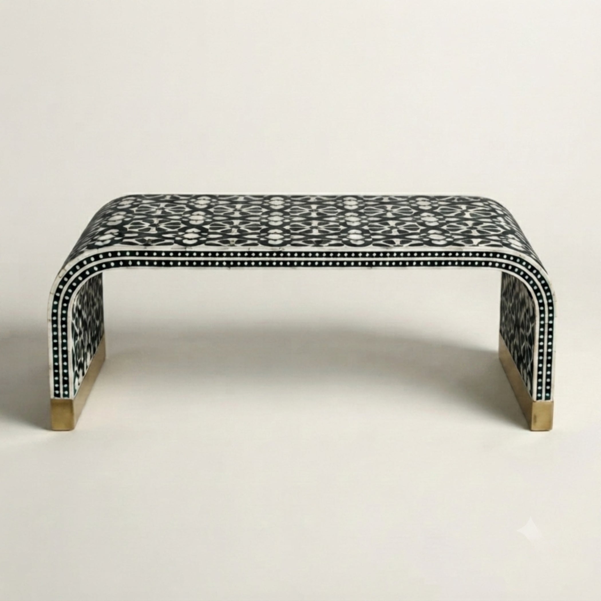 A low curved bench with a black and white geometric bone-inlay pattern and gold-toned metal feet, photographed against a plain light gray studio background.